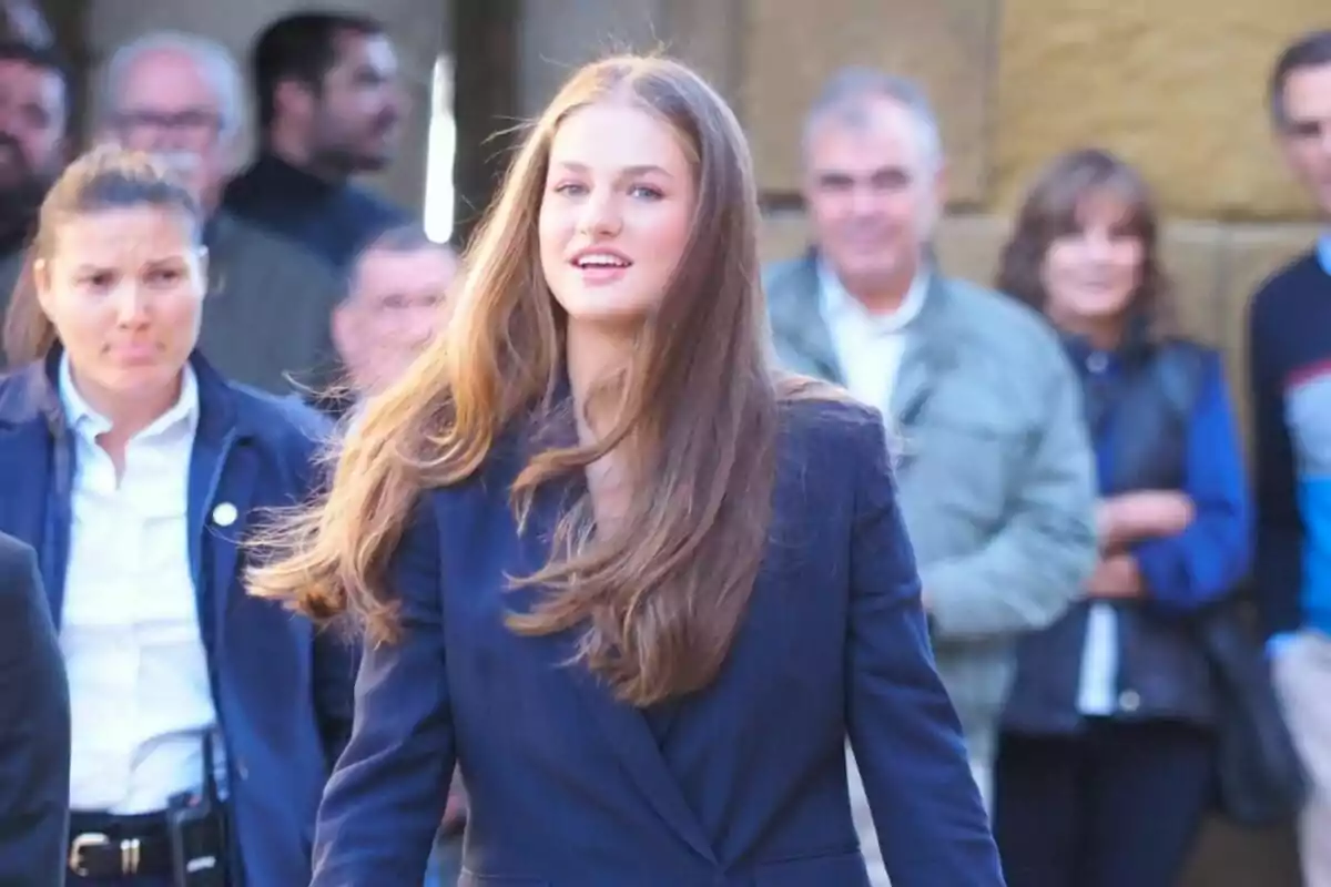 Jove de cabell llarg i llis amb jaqueta blava caminant a l’aire lliure mentre diverses persones l’observen al fons Jove de cabell llarg i llis amb jaqueta blava caminant a l’aire lliure mentre diverses persones l’observen al fons