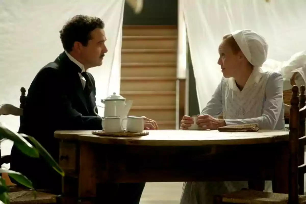 Un home i una dona vestits amb roba antiga conversen asseguts davant d’una taula de fusta amb una tetera i tasses.