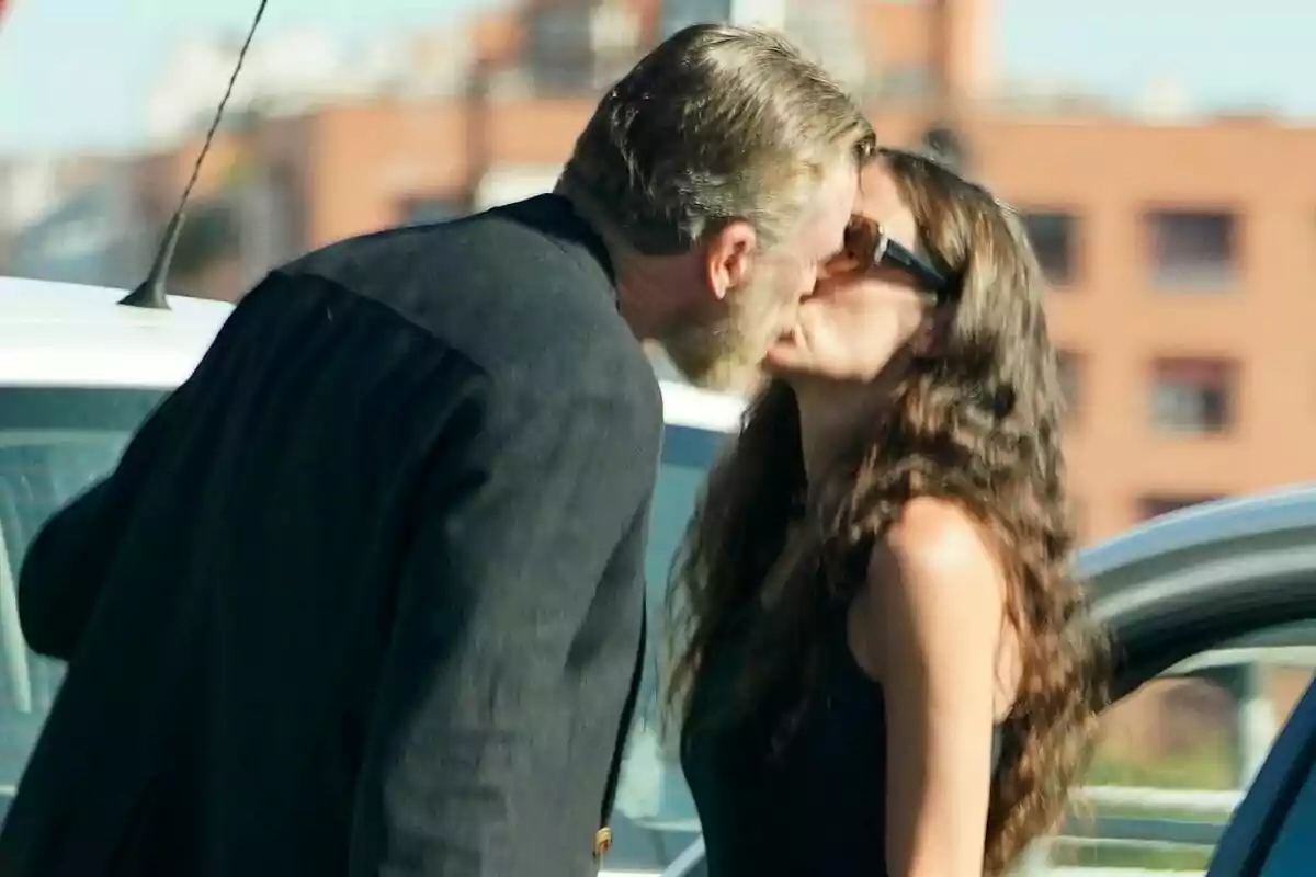 Parella besant-se a l’aire lliure al costat d’un cotxe amb edificis desenfocats al fons Parella besant-se a l’aire lliure al costat d’un cotxe amb edificis desenfocats al fons