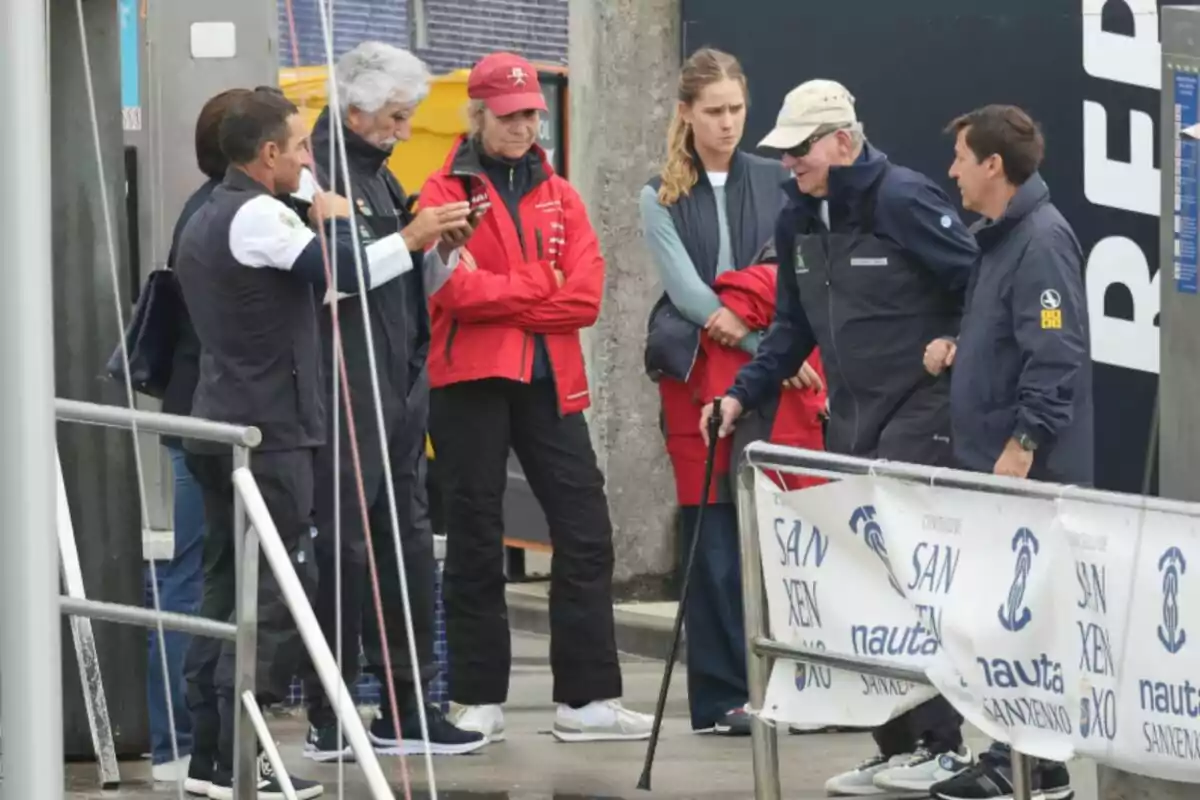Un grup de persones vestides amb roba esportiva i abrics conversen i observen alguna cosa en un port al costat d’una barana i una lona amb logotips d’un esdeveniment nàutic.