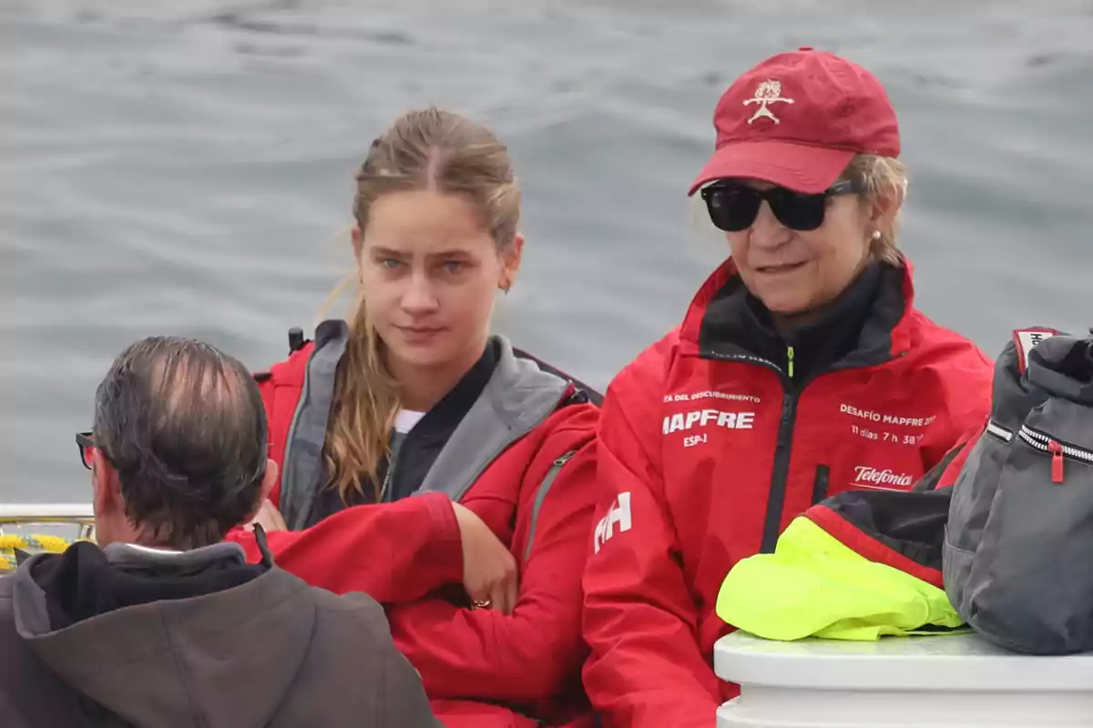 Irene Urdangarin, la infanta Elena i una altra persona amb jaquetes vermelles i una gorra estan assegudes en una barca sobre l’aigua, una d’elles porta ulleres de sol i una altra mira al davant amb expressió seriosa. Irene Urdangarin, la infanta Elena i una altra persona amb jaquetes vermelles i una gorra estan assegudes en una barca sobre l’aigua, una d’elles porta ulleres de sol i una altra mira al davant amb expressió seriosa.