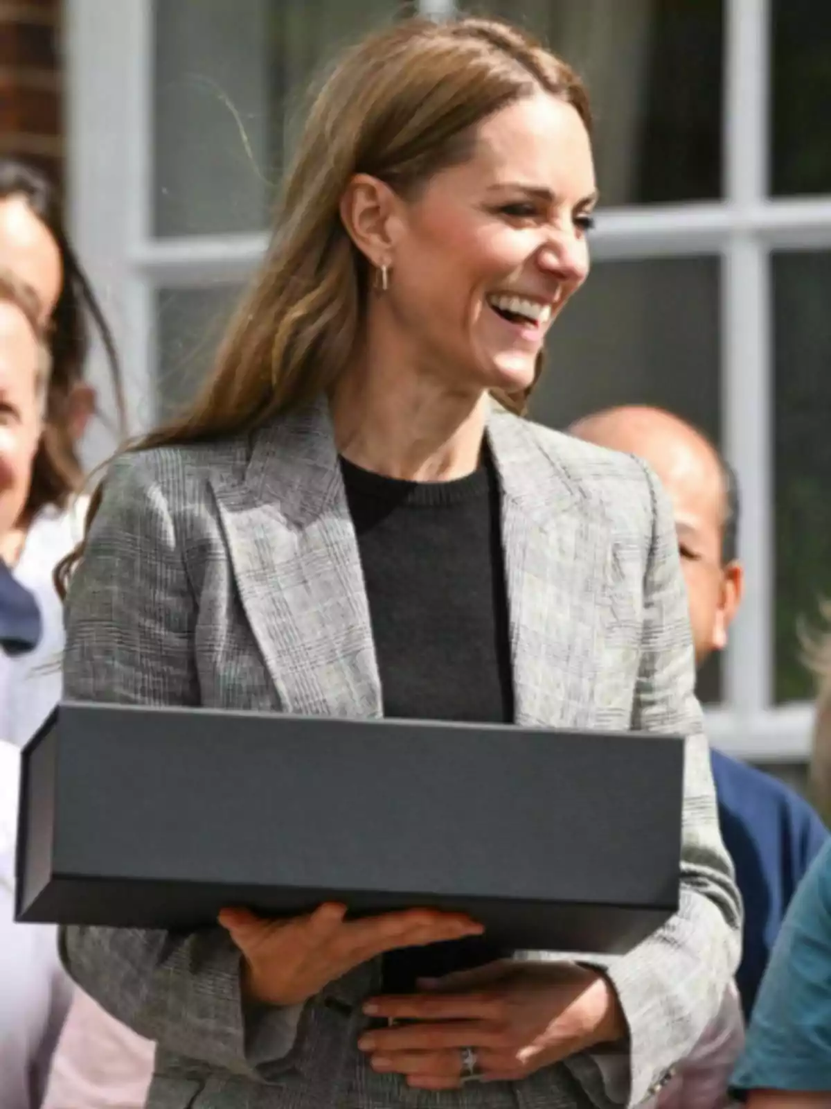 Kate Middleton somrient amb una jaqueta grisa sosté una caixa negra rectangular mentre està envoltada d’altres persones en un ambient exterior.
