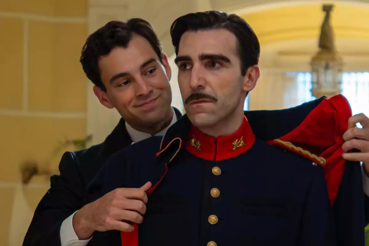 Curro i Lorenzo, elegantment vestits, l’un ajuda l’altre a posar-se un abric militar blau amb detalls daurats i coll vermell en un ambient interior il·luminat. Captura de 'La Promesa' Curro i Lorenzo, elegantment vestits, l’un ajuda l’altre a posar-se un abric militar blau amb detalls daurats i coll vermell en un ambient interior il·luminat. Captura de 'La Promesa'