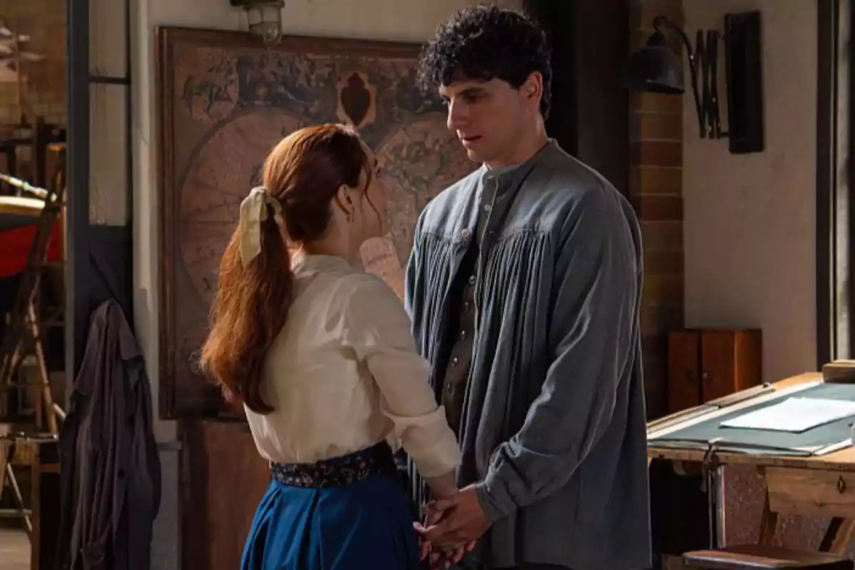 Enora i Toño es miren als ulls i es donen les mans en una habitació amb decoració antiga i un gran mapa a la paret. Captura de 'La Promesa'