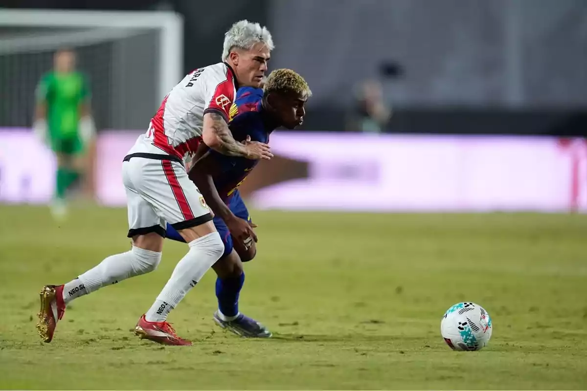 Dos futbolistes disputen la pilota durant un partit en un estadi, un amb uniforme blanc i vermell i l’altre amb uniforme blau fosc. Dos futbolistes disputen la pilota durant un partit en un estadi, un amb uniforme blanc i vermell i l’altre amb uniforme blau fosc.