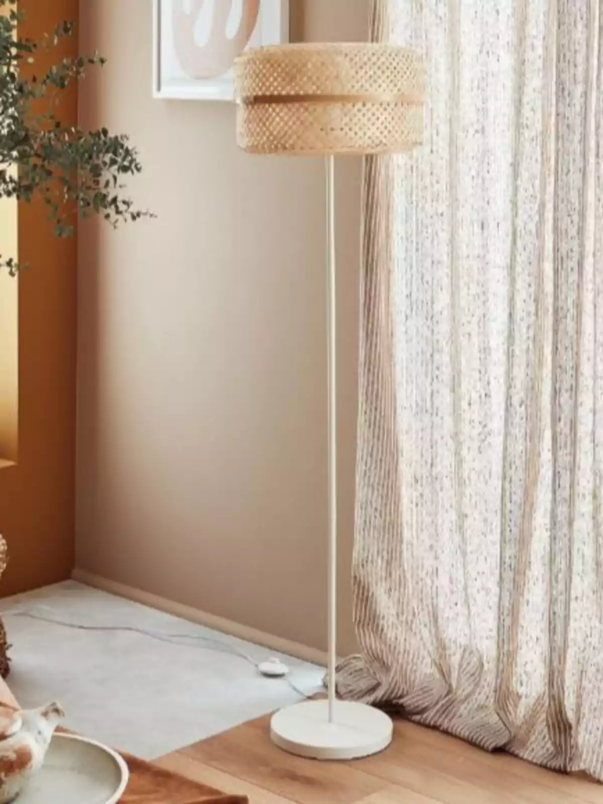 Làmpada de peu amb base i estructura blanca i pantalla de vímet en una sala decorada en tons neutres al costat d’una cortina clara