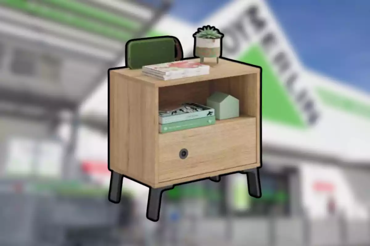 Tauleta de nit de fusta clara amb potes negres, decorada amb llibres, una planta petita i objectes a sobre i dins del moble, amb un fons desenfocat d’una botiga.