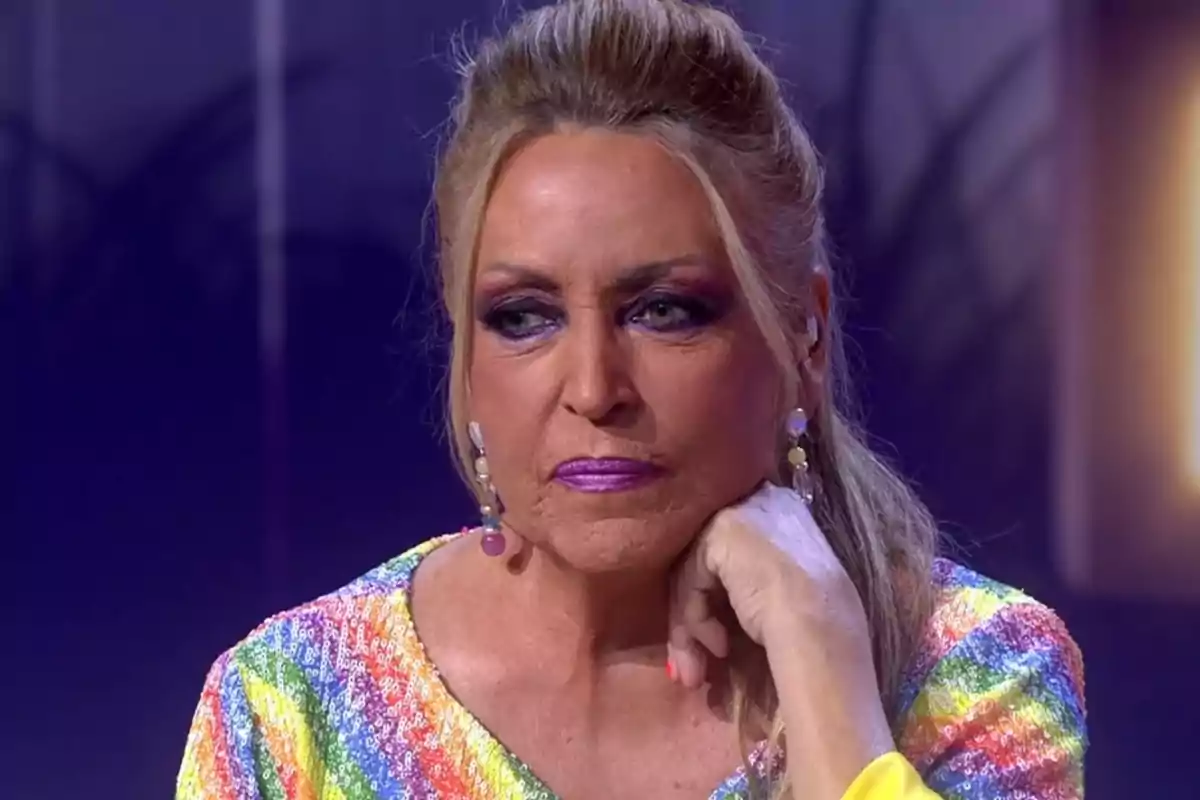Lydia Lozano, con maquillaje llamativo y expresión seria, apoyando la barbilla en su mano y vistiendo una blusa de lentejuelas multicolor