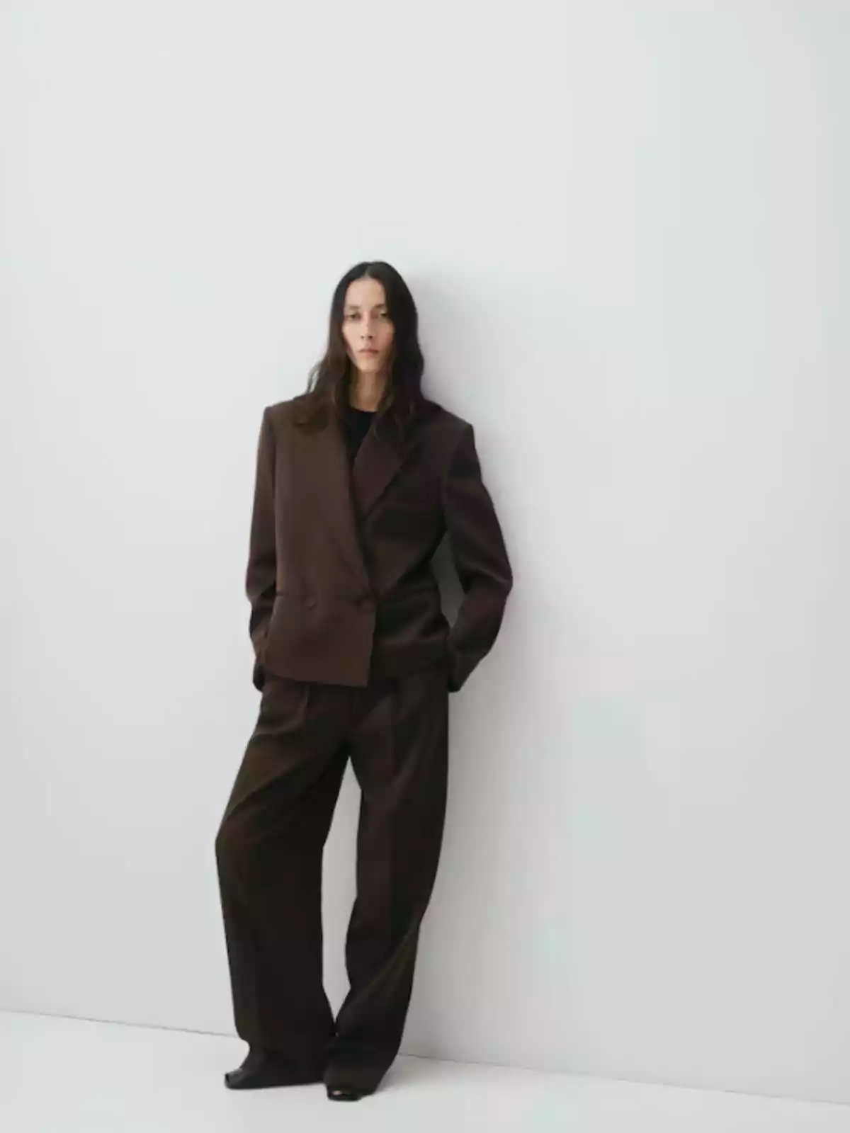 Persona de cabello largo y suelto vestida con un traje marrón de chaqueta y pantalón ancho, posando de pie junto a una pared blanca Persona de cabello largo y suelto vestida con un traje marrón de chaqueta y pantalón ancho, posando de pie junto a una pared blanca