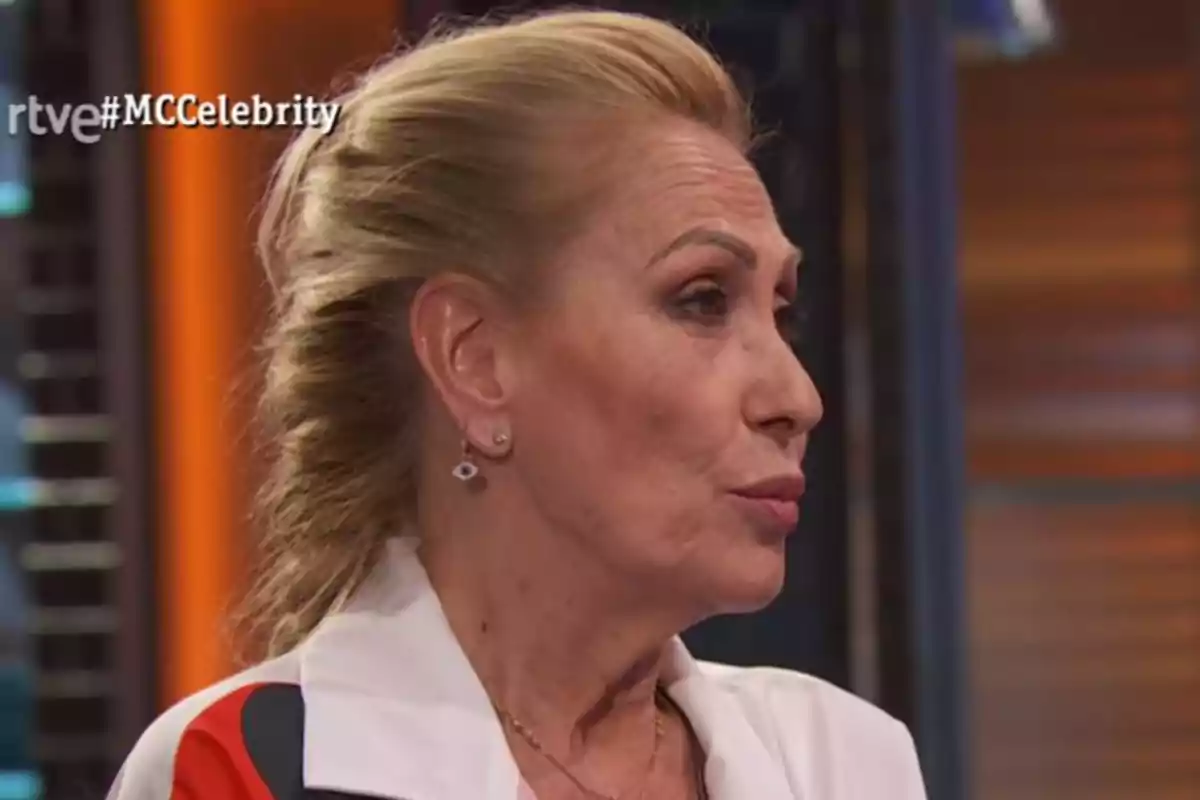 Rosa Benito de perfil con el cabello recogido y una chaqueta blanca en un set de televisión con el hashtag MCCelebrity en la esquina superior izquierda Rosa Benito de perfil con el cabello recogido y una chaqueta blanca en un set de televisión con el hashtag MCCelebrity en la esquina superior izquierda