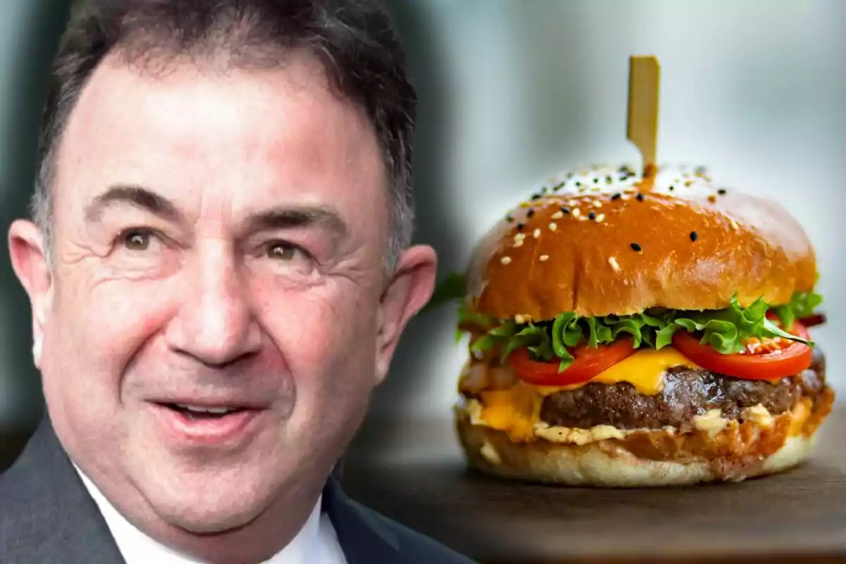 Martín Berasategui amb vestit somriu mentre una hamburguesa amb formatge, enciam i tomàquet és al seu costat sobre una taula. Martín Berasategui amb vestit somriu mentre una hamburguesa amb formatge, enciam i tomàquet és al seu costat sobre una taula.