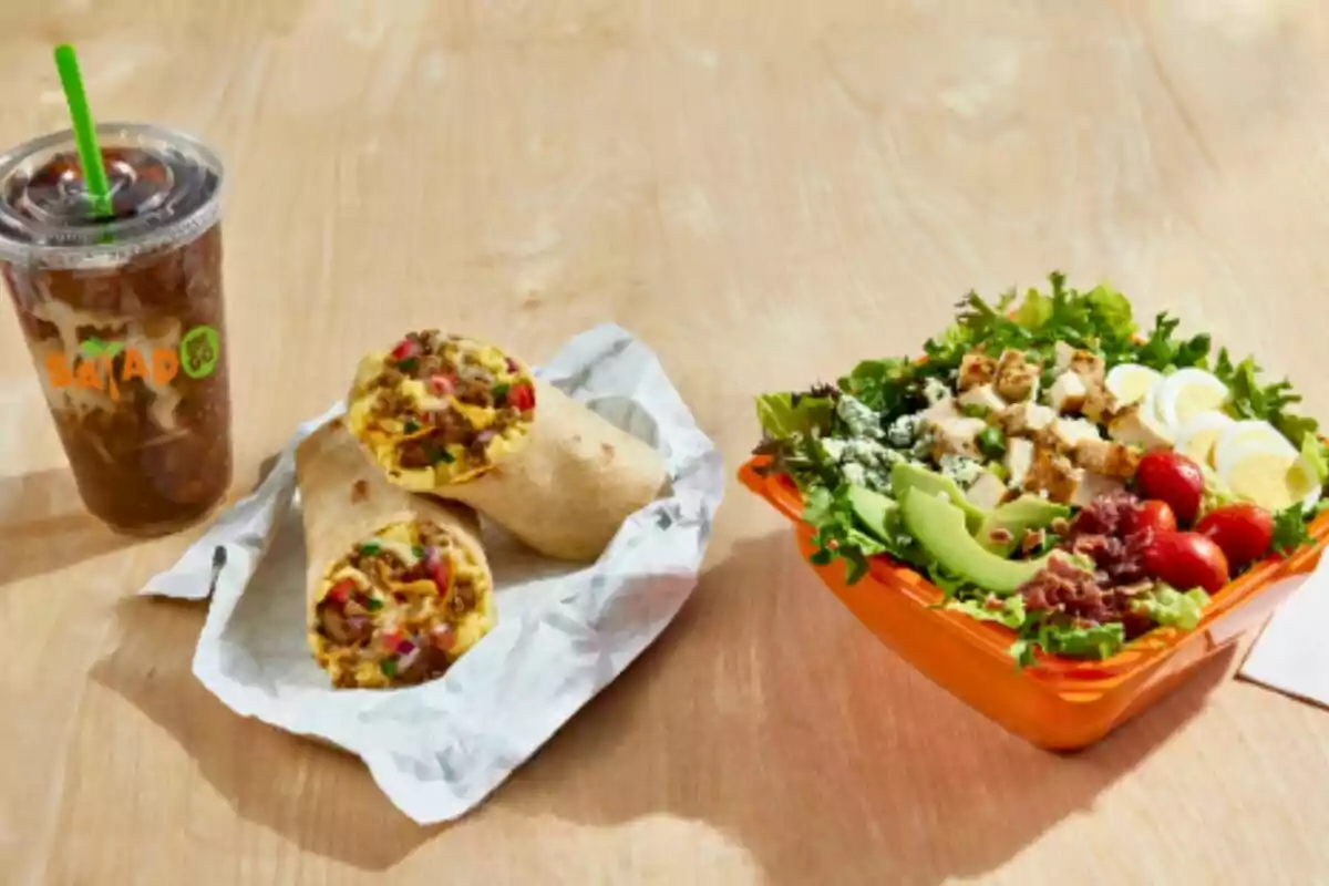 Cold drink in a plastic cup next to a burrito cut in half and a salad with chicken, egg, avocado, tomatoes, and bacon on a wooden table