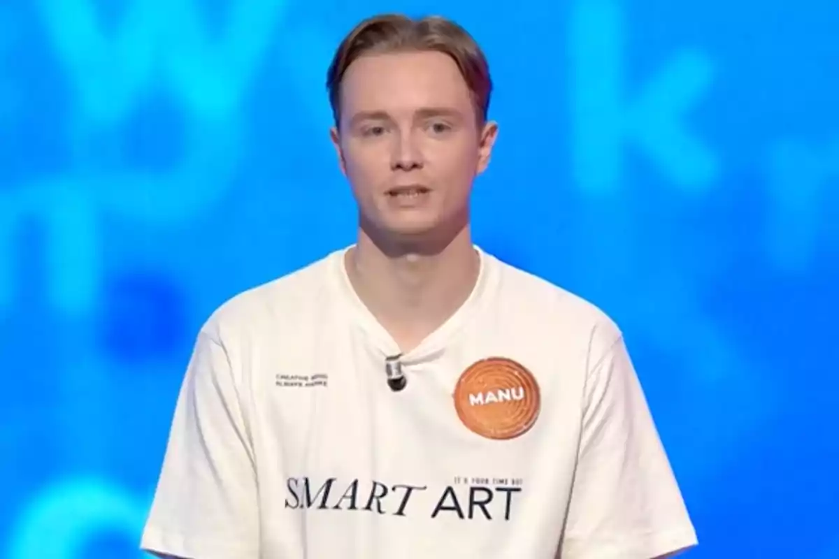 Manu amb samarreta blanca que té el text SMART ART i una etiqueta taronja amb el nom Manu sobre un fons blau. Captura de 'Pasapalabra' Manu amb samarreta blanca que té el text SMART ART i una etiqueta taronja amb el nom Manu sobre un fons blau. Captura de 'Pasapalabra'