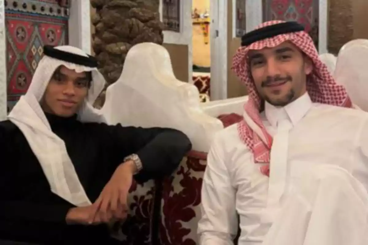 Alexandre Grimaldi and another man in traditional Arab clothing are sitting on a decorated sofa; both are smiling and appear relaxed in an indoor setting with colorful details on the walls.