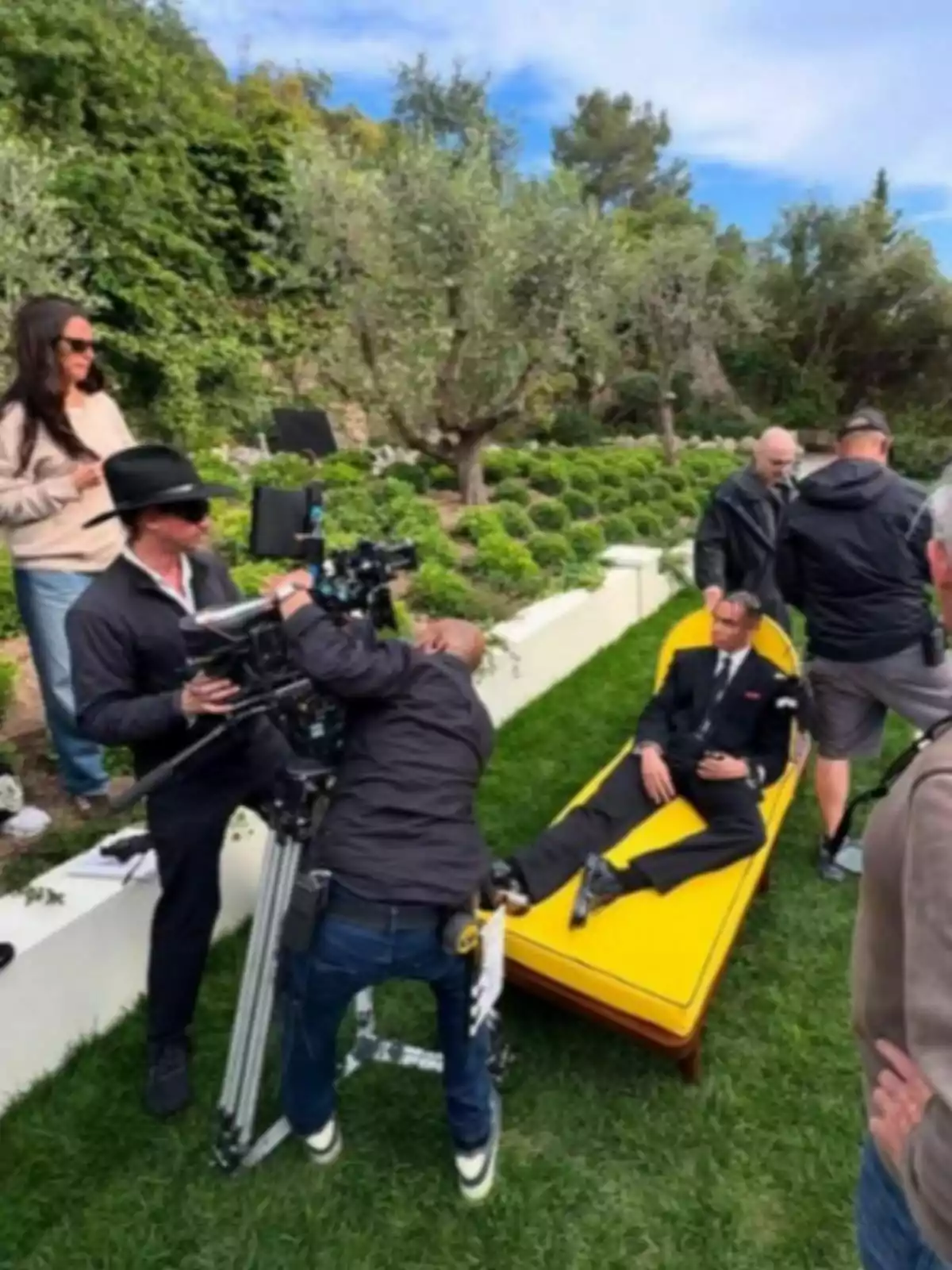 A film crew records Alexandre Grimaldi, dressed in a suit, lying on a yellow divan in a garden surrounded by greenery.