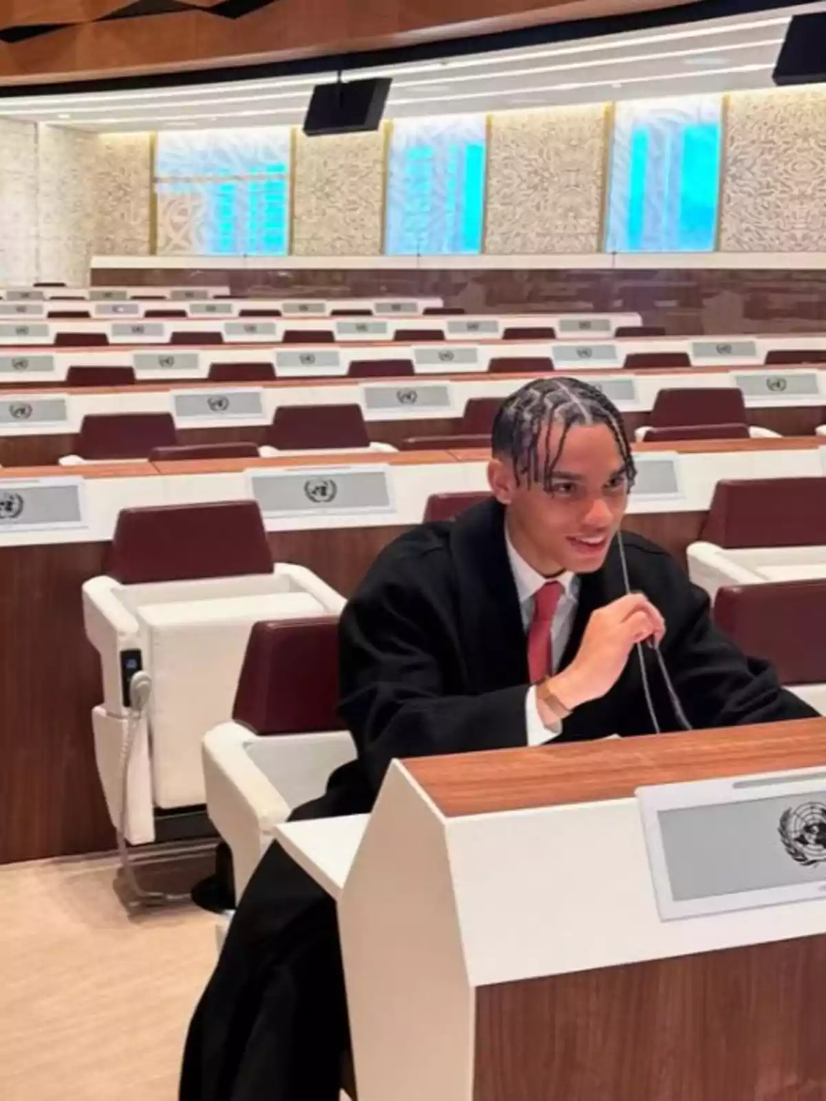 Alexandre Grimaldi in a suit and red tie speaking into a microphone inside a conference room with empty seats and UN emblems