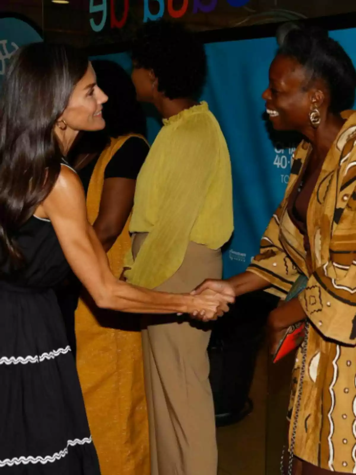 La reina Letícia i una altra dona somrients es donen la mà mentre dues altres persones conversen al fons. La reina Letícia i una altra dona somrients es donen la mà mentre dues altres persones conversen al fons.