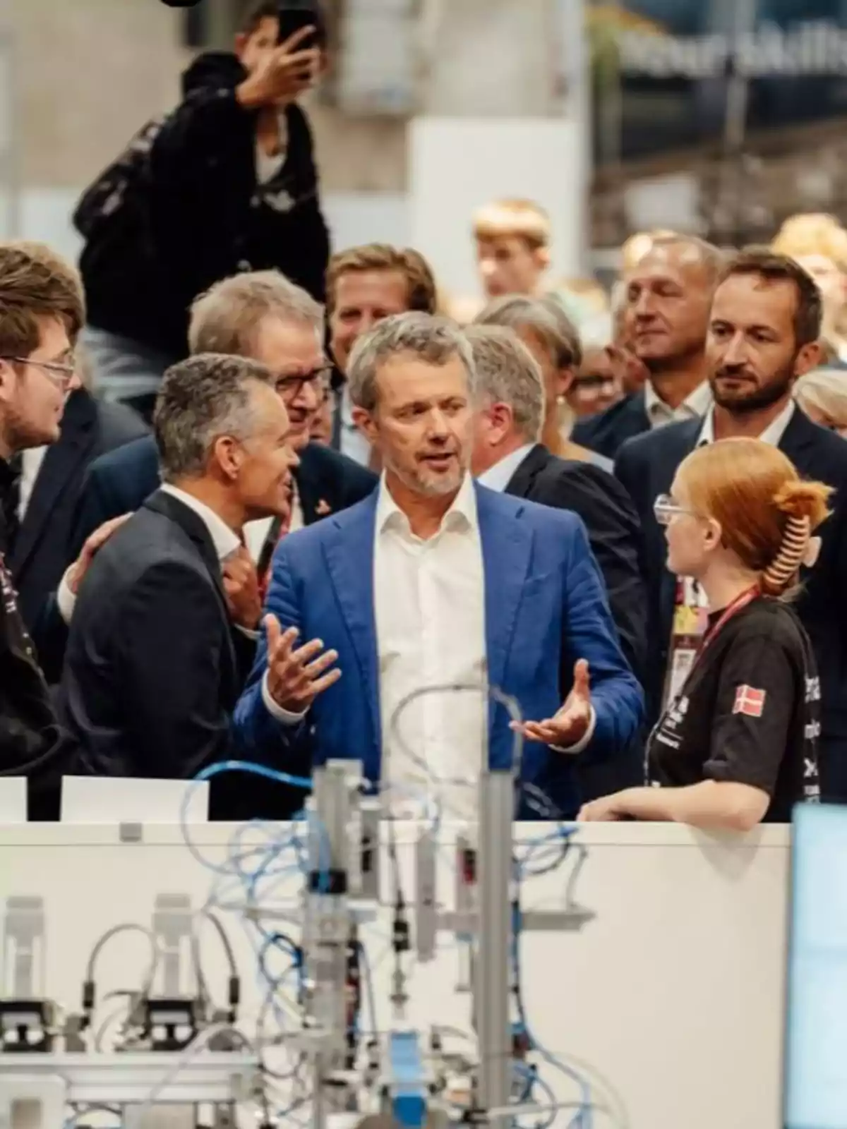 Federico de Dinamarca de traje azul conversa animadamente con una joven frente a un grupo de personas en un entorno tecnológico