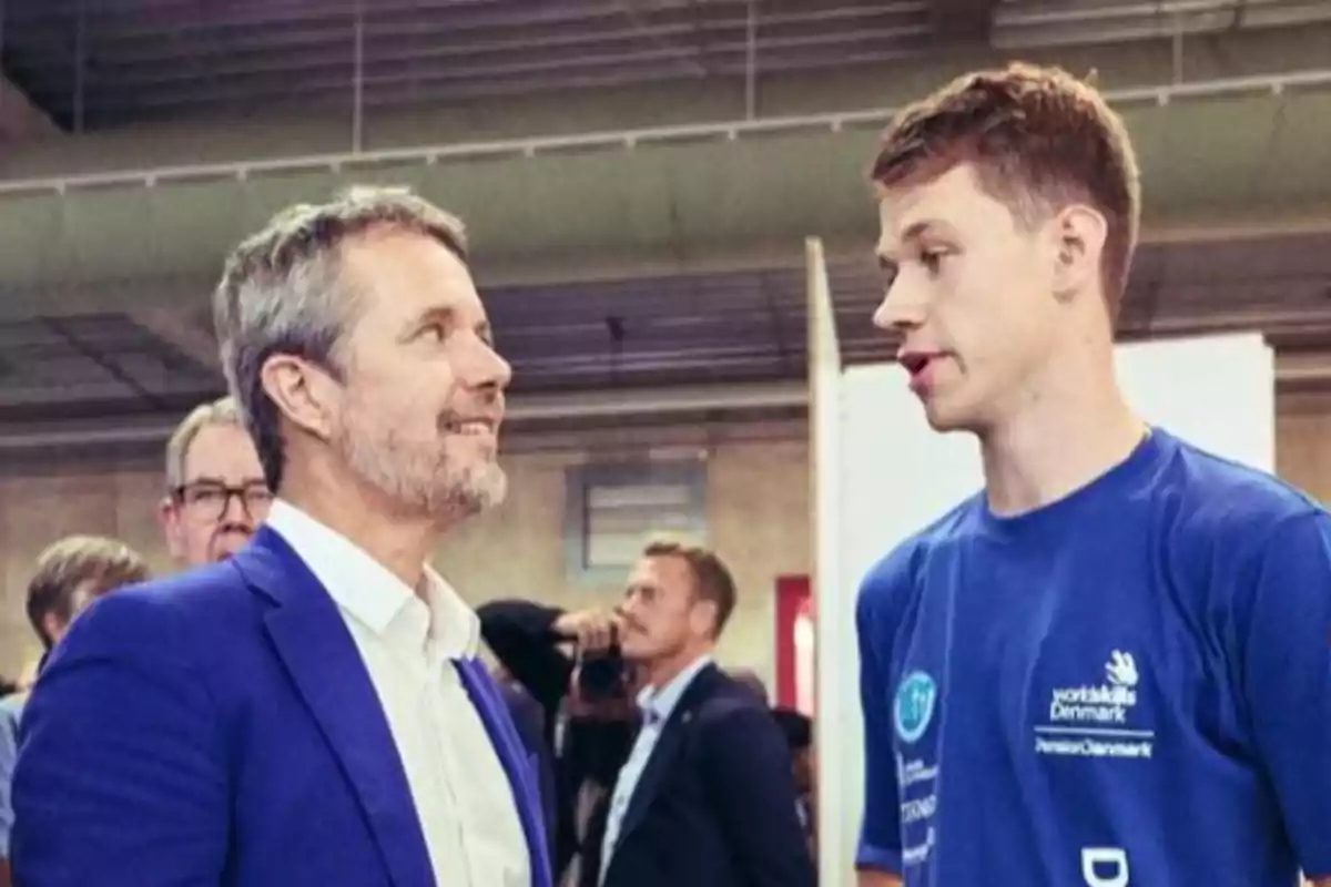 Federico de Dinamarca y otro hombre conversan en un espacio interior mientras otras personas se ven al fondo