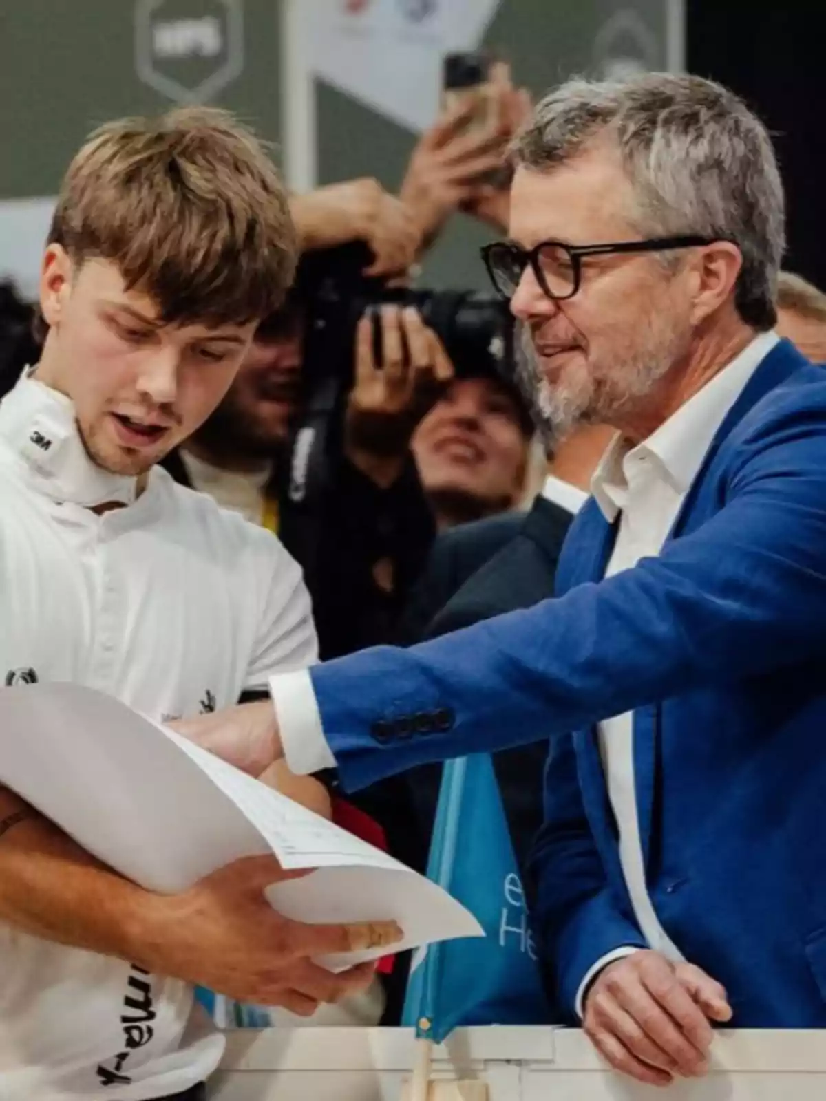 Federico de Dinamarca y otro hombre conversan mientras revisan unos papeles en un evento, uno de ellos lleva gafas y saco azul, el otro viste camiseta blanca y sostiene los documentos.