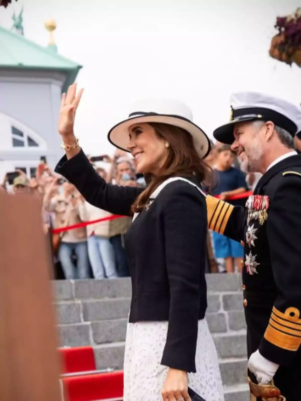 Mary de Dinamarca amb barret blanc i vestit clar saluda somrient al costat de Frederic de Dinamarca amb uniforme militar mentre una multitud observa al fons. Mary de Dinamarca amb barret blanc i vestit clar saluda somrient al costat de Frederic de Dinamarca amb uniforme militar mentre una multitud observa al fons.