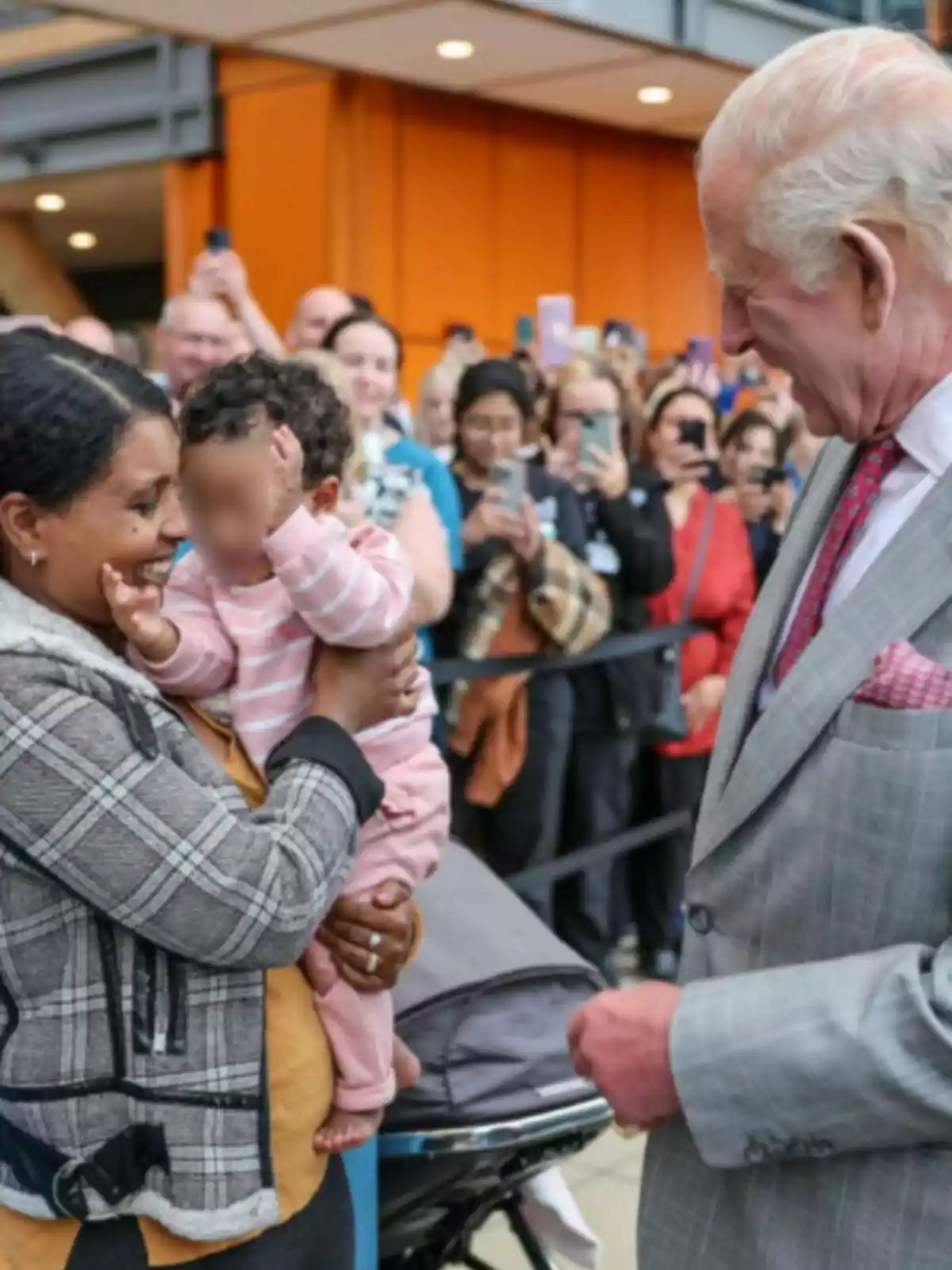 Carlos III elegantemente vestido sonríe mientras conversa con una mujer que sostiene a un bebé en brazos, rodeados de una multitud que toma fotos con sus teléfonos móviles. Carlos III elegantemente vestido sonríe mientras conversa con una mujer que sostiene a un bebé en brazos, rodeados de una multitud que toma fotos con sus teléfonos móviles.