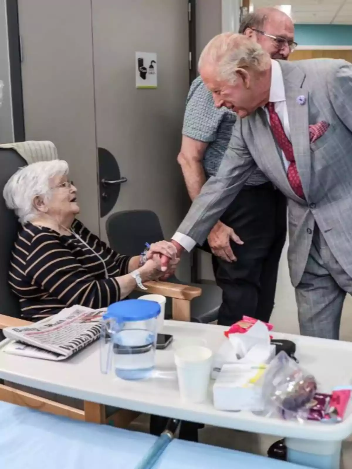 Carlos III elegantemente vestido saluda de la mano a una mujer mayor sentada en una silla dentro de una habitación, mientras otra persona los observa y una mesa con objetos está en primer plano. Carlos III elegantemente vestido saluda de la mano a una mujer mayor sentada en una silla dentro de una habitación, mientras otra persona los observa y una mesa con objetos está en primer plano.