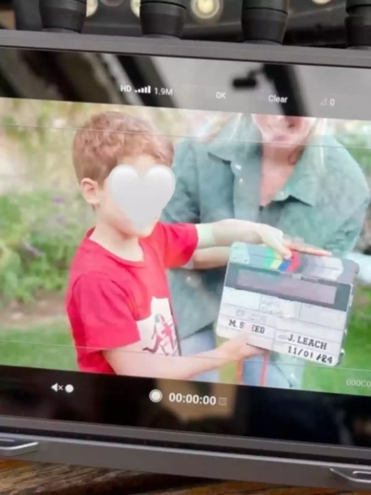 Un nen amb samarreta vermella sosté una claqueta de cinema mentre una persona adulta l’acompanya en un entorn a l’aire lliure; tots dos apareixen a la pantalla d’un monitor de gravació. Un nen amb samarreta vermella sosté una claqueta de cinema mentre una persona adulta l’acompanya en un entorn a l’aire lliure; tots dos apareixen a la pantalla d’un monitor de gravació.