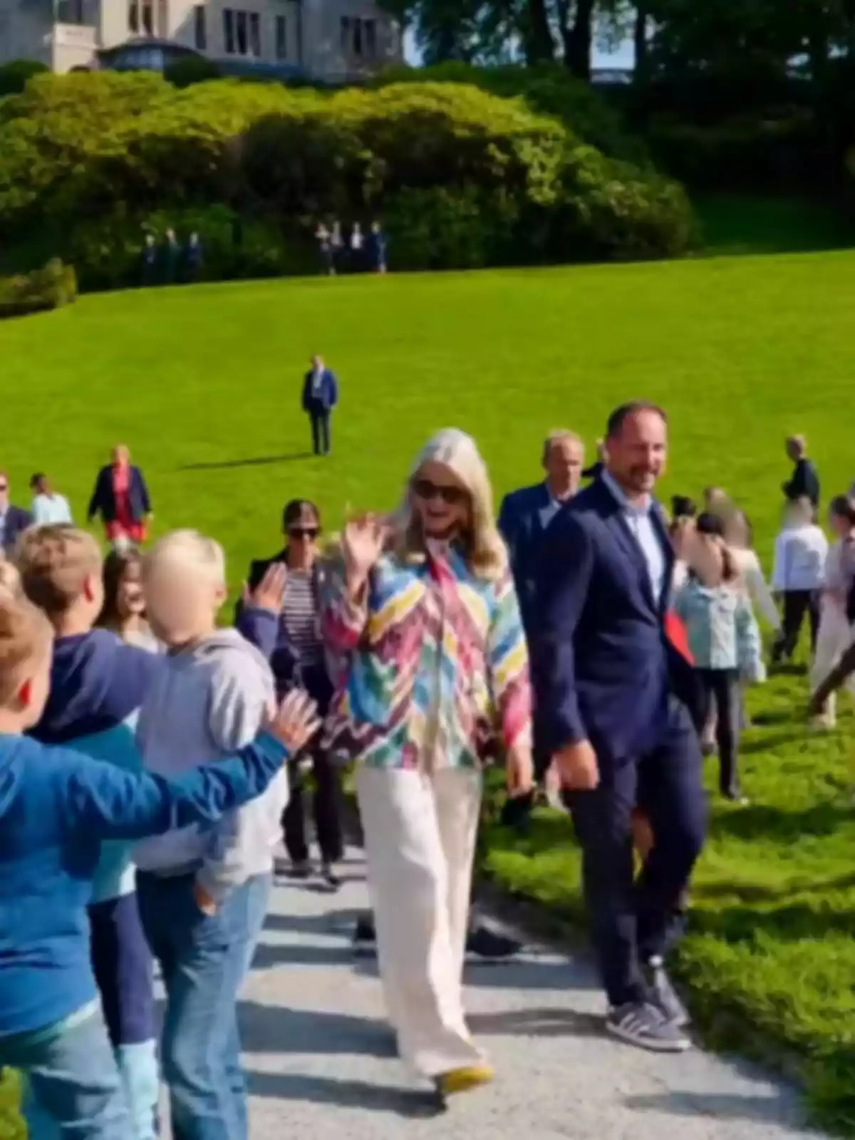 Mette-Marit con gafas de sol y ropa colorida camina por un sendero saludando a un grupo de niños mientras Haakon con traje la acompaña en un entorno de césped verde y personas al fondo Mette-Marit con gafas de sol y ropa colorida camina por un sendero saludando a un grupo de niños mientras Haakon con traje la acompaña en un entorno de césped verde y personas al fondo