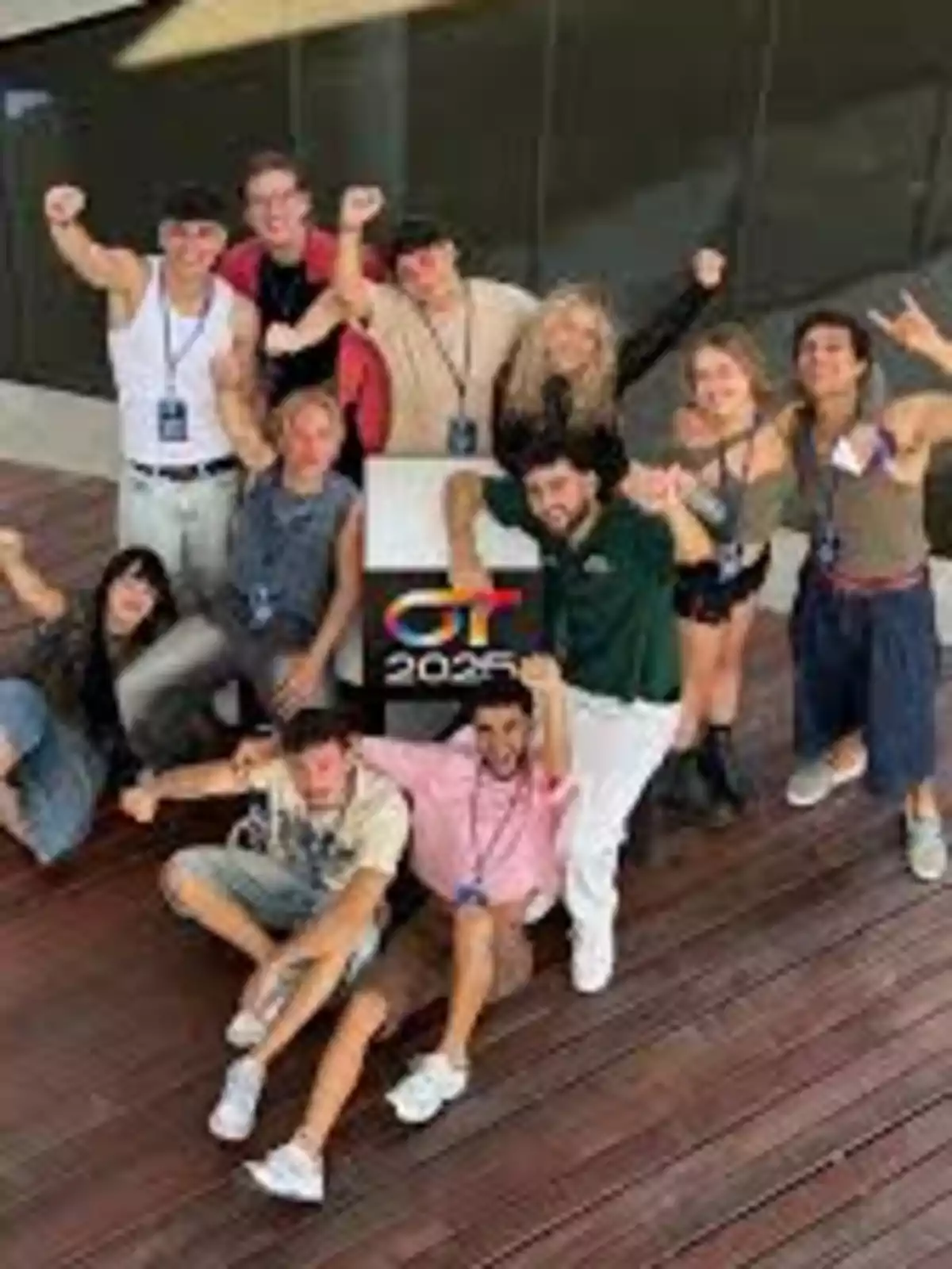 Un grupo de jóvenes sonríe y celebra mientras posa en círculo sobre un piso de madera, algunos levantan el brazo y en el centro hay un cartel con el logo de OT 2023.