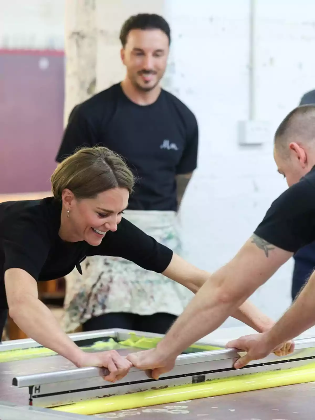 Kate Middleton somrient i un home col·laboren en una activitat de serigrafia mentre una altra persona els observa al fons.