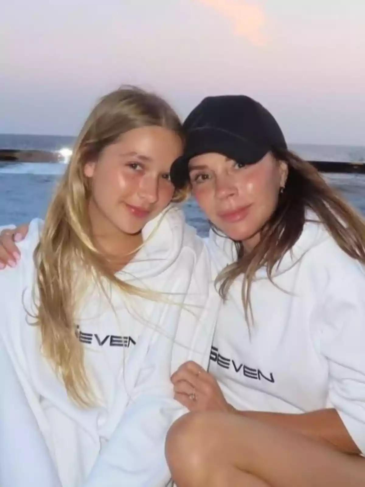 Harper y Victoria Beckham con sudaderas blancas posan juntas frente al mar al atardecer Harper y Victoria Beckham con sudaderas blancas posan juntas frente al mar al atardecer
