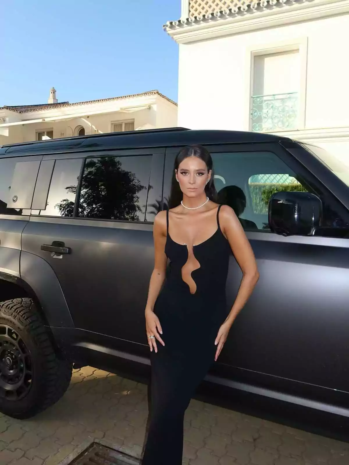 Victoria Federica con vestido negro elegante posando frente a una camioneta negra estacionada junto a una casa de lujo en un día soleado