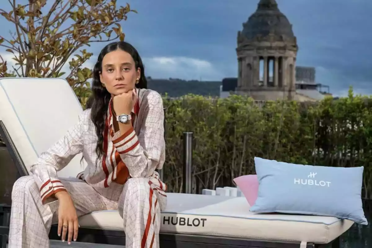 Victoria Federica sentada en un diván al aire libre con ropa elegante y reloj, al fondo vegetación y una estructura arquitectónica antigua