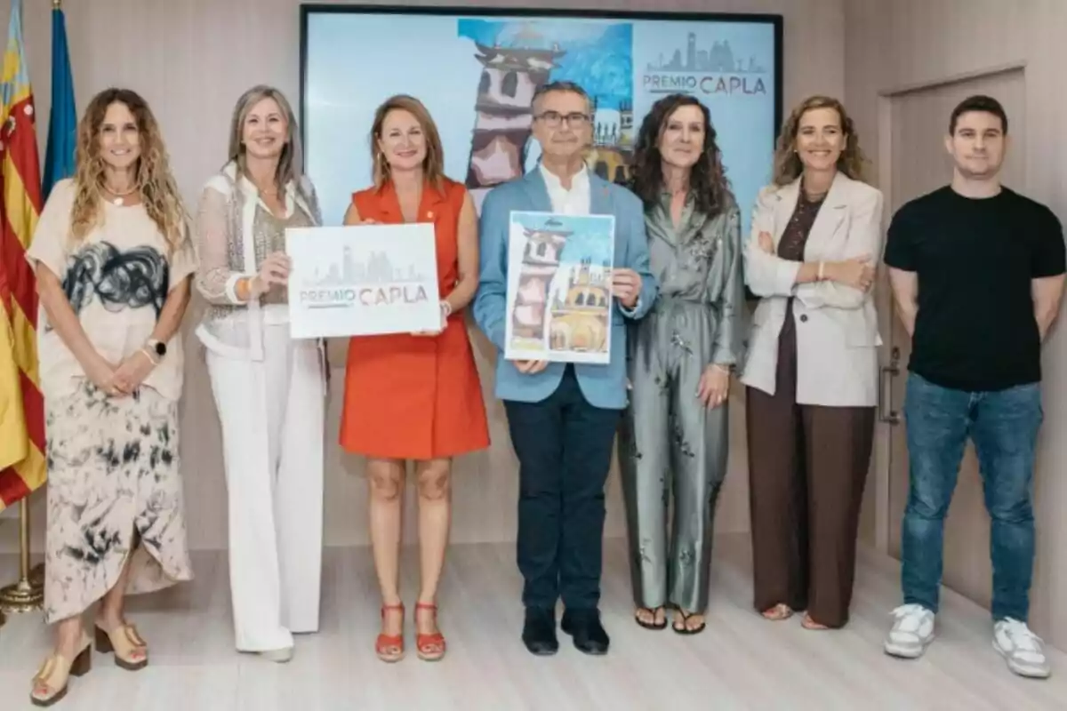 Set persones posen en una sala al costat d’una pantalla i banderes, dues d’elles sostenen cartells relacionats amb un premi anomenat CAPLA. Set persones posen en una sala al costat d’una pantalla i banderes, dues d’elles sostenen cartells relacionats amb un premi anomenat CAPLA.