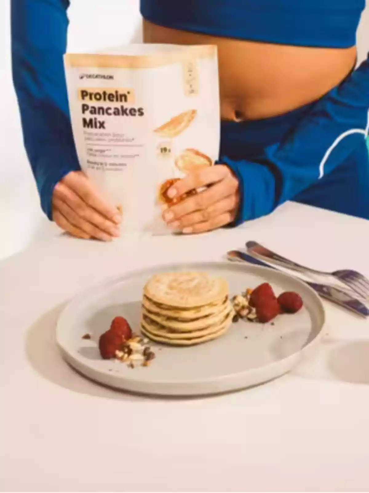 Persona amb roba esportiva blava sosté una bossa de barreja per a pancakes de proteïna sobre una taula on hi ha un plat amb una torre de pancakes, maduixes i fruits secs.