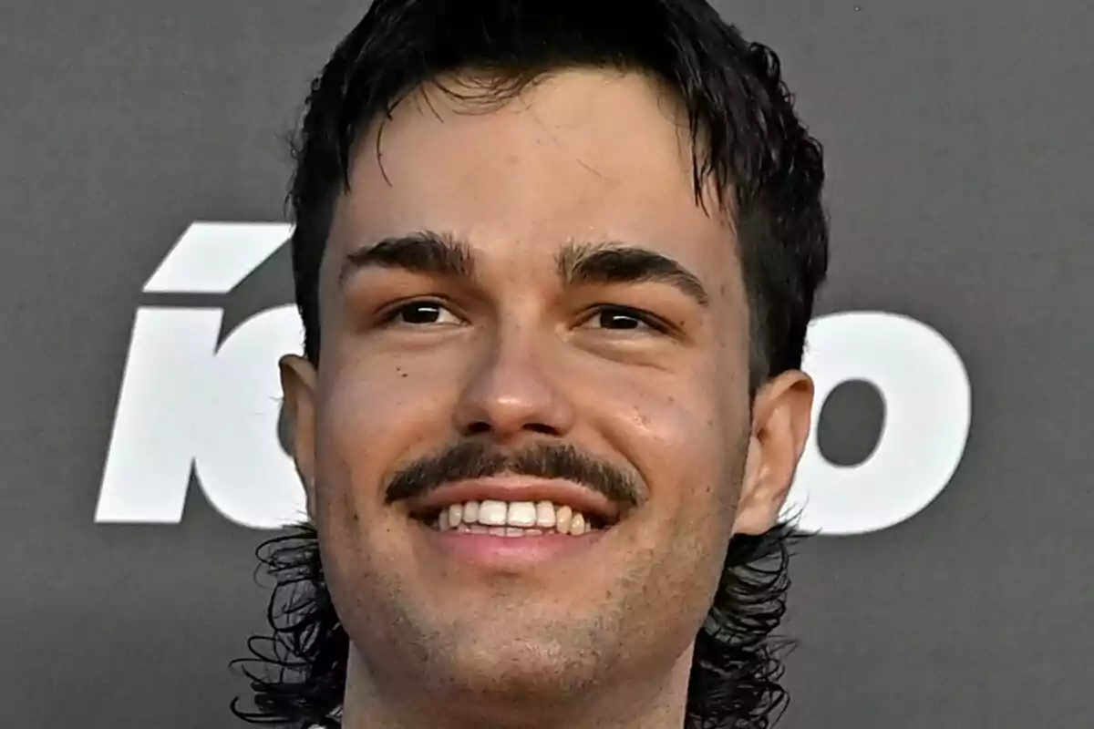 Primer plano de Peldanyos sonriendo frente a un fondo gris con letras blancas desenfocadas.