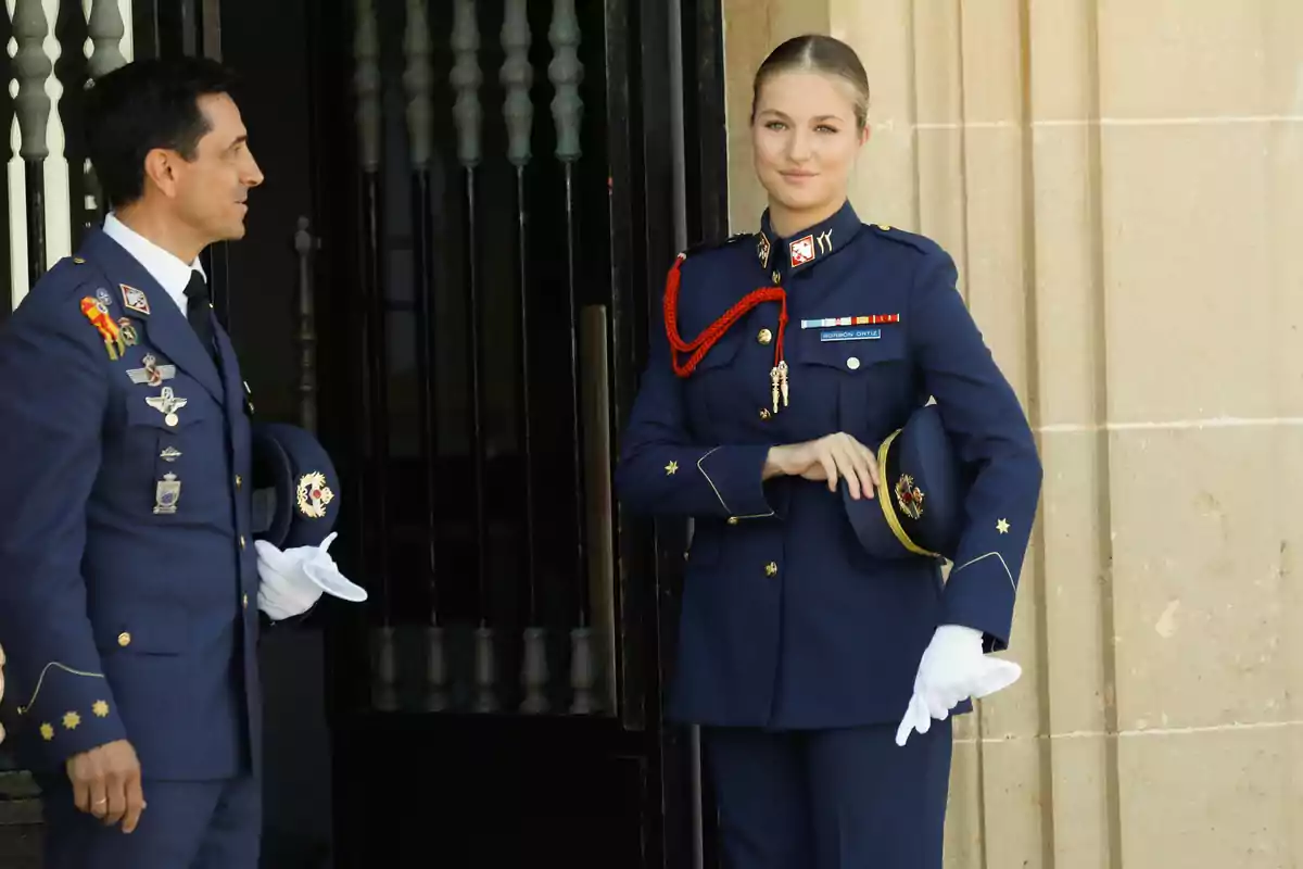 La princesa Leonor amb uniforme militar blau posen a l’entrada d’un edifici de pedra, una d’elles sosté una gorra i porta guants blancs.