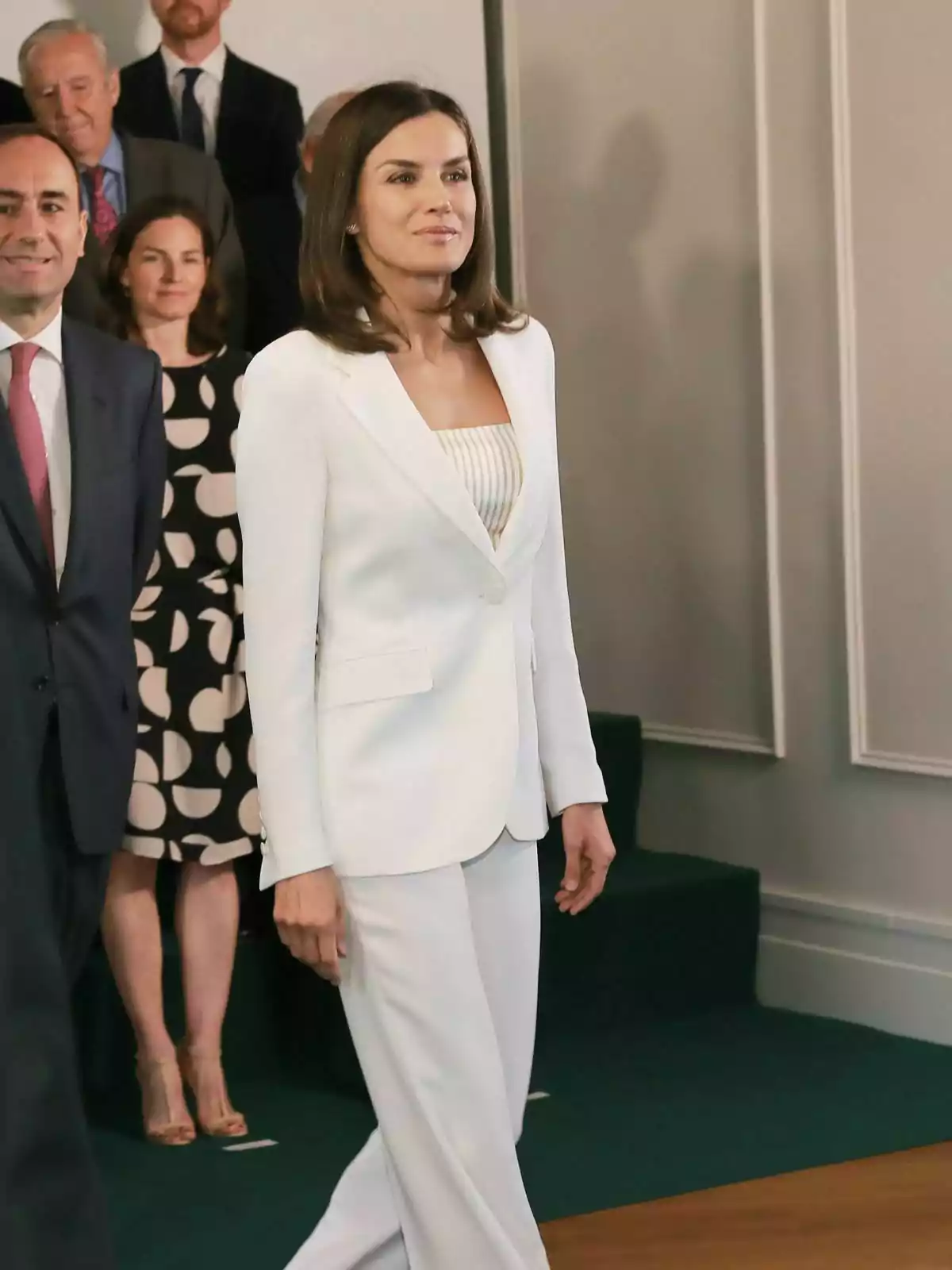 Letícia Ortiz amb vestit blanc caminant en un esdeveniment formal amb diverses persones al fons. Letícia Ortiz amb vestit blanc caminant en un esdeveniment formal amb diverses persones al fons.