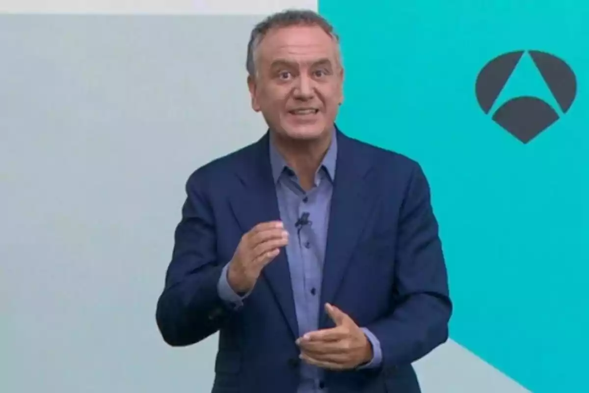 Plano medio de Roberto Brasero hablando frente a una pared de fondo azul claro con un logotipo geométrico oscuro a la derecha.