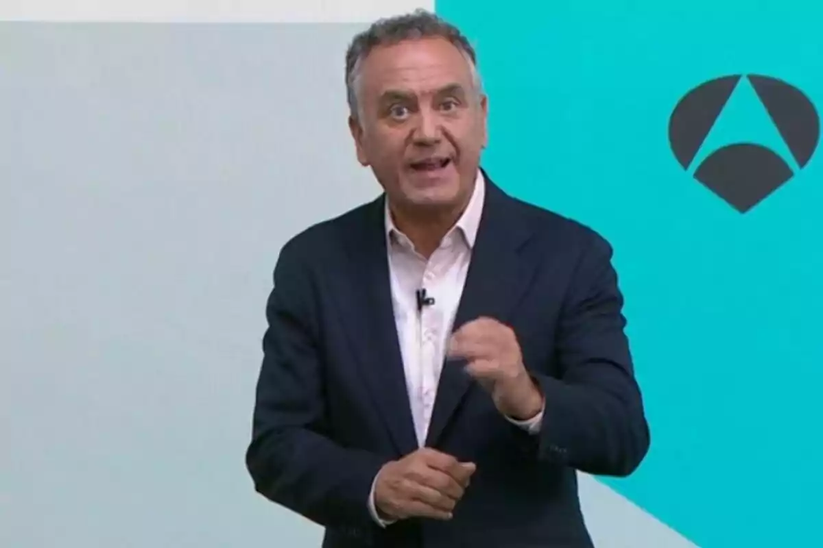 Plano medio de Roberto Brasero vestido con traje oscuro y camisa blanca hablando frente a un fondo azul con un logotipo geométrico, que es de Antena 3. Plano medio de Roberto Brasero vestido con traje oscuro y camisa blanca hablando frente a un fondo azul con un logotipo geométrico, que es de Antena 3.