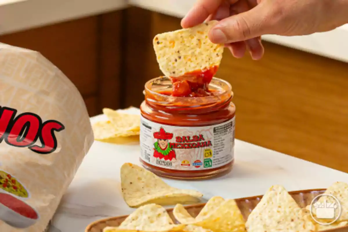 Una mà sosté un totopo submergit en un pot de salsa mexicana al costat d’una bossa de nachos i diversos totopos sobre la taula.
