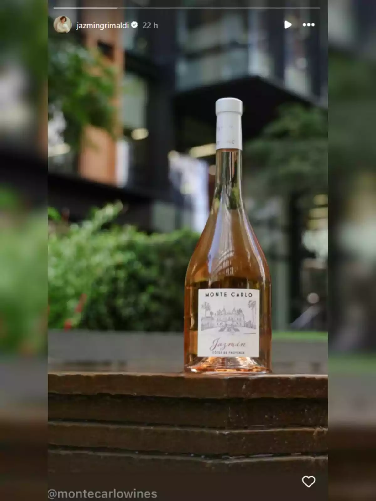 Ampolla de vi rosat Monte Carlo Jasmin col·locada sobre una superfície de fusta amb un fons de jardí i edificis moderns desenfocats Ampolla de vi rosat Monte Carlo Jasmin col·locada sobre una superfície de fusta amb un fons de jardí i edificis moderns desenfocats