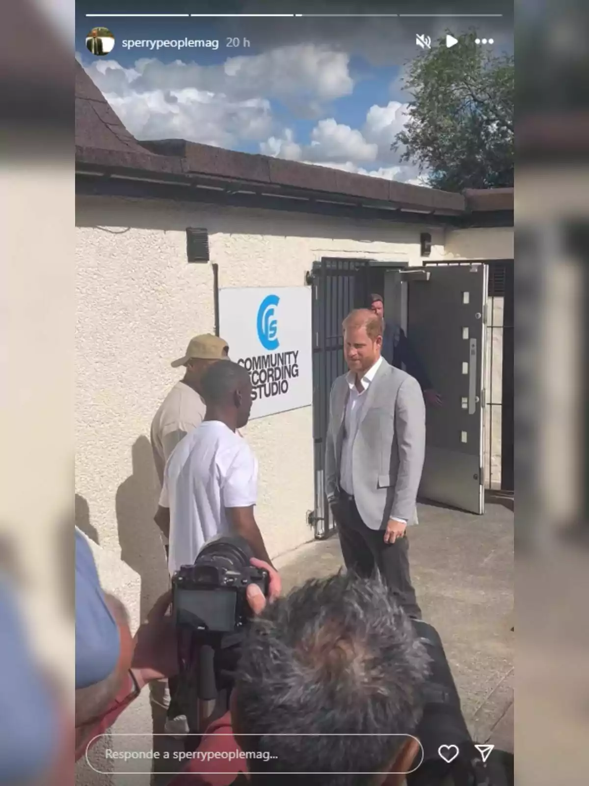El príncep Harry amb americana grisa conversa amb altres persones davant d’un edifici amb un rètol que diu Community Recording Studio mentre diverses càmeres el fotografien.
