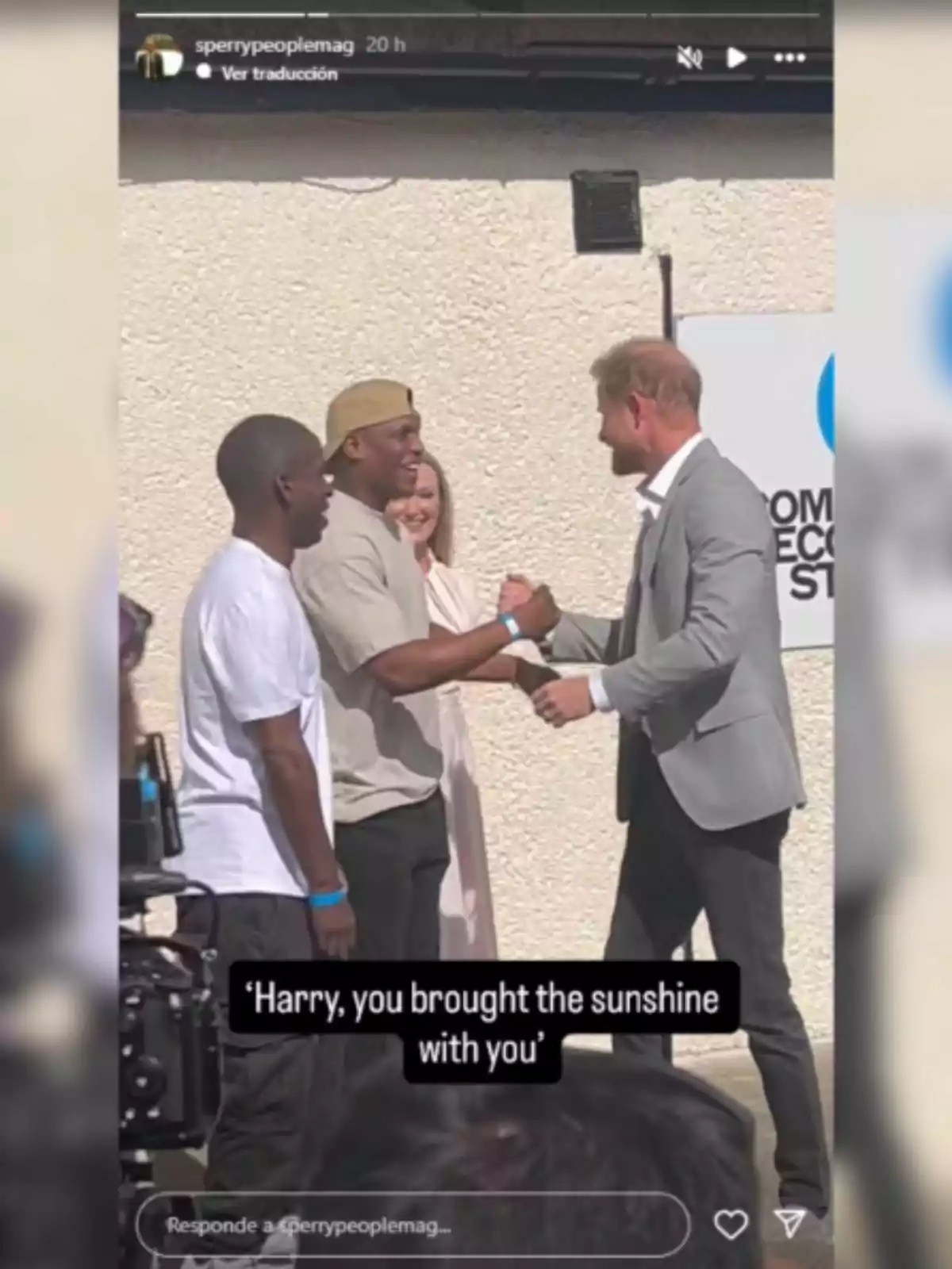 El príncep Harry, dos homes i una dona somriuen mentre dos d’ells es saluden de mà en un ambient assolellat, amb una càmera gravant l’escena i un rètol parcialment visible al fons.