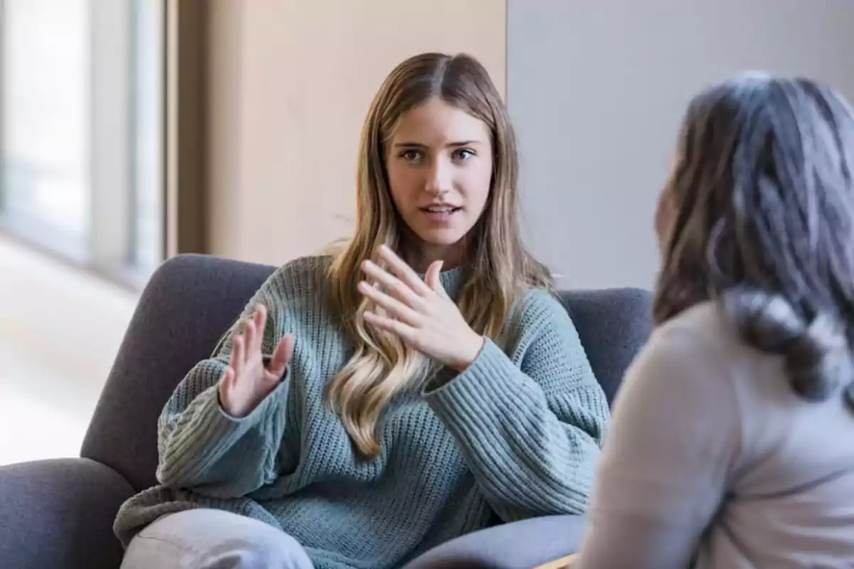 Dos mujeres sentadas conversando en una sala iluminada mientras una de ellas gesticula con las manos Dos mujeres sentadas conversando en una sala iluminada mientras una de ellas gesticula con las manos
