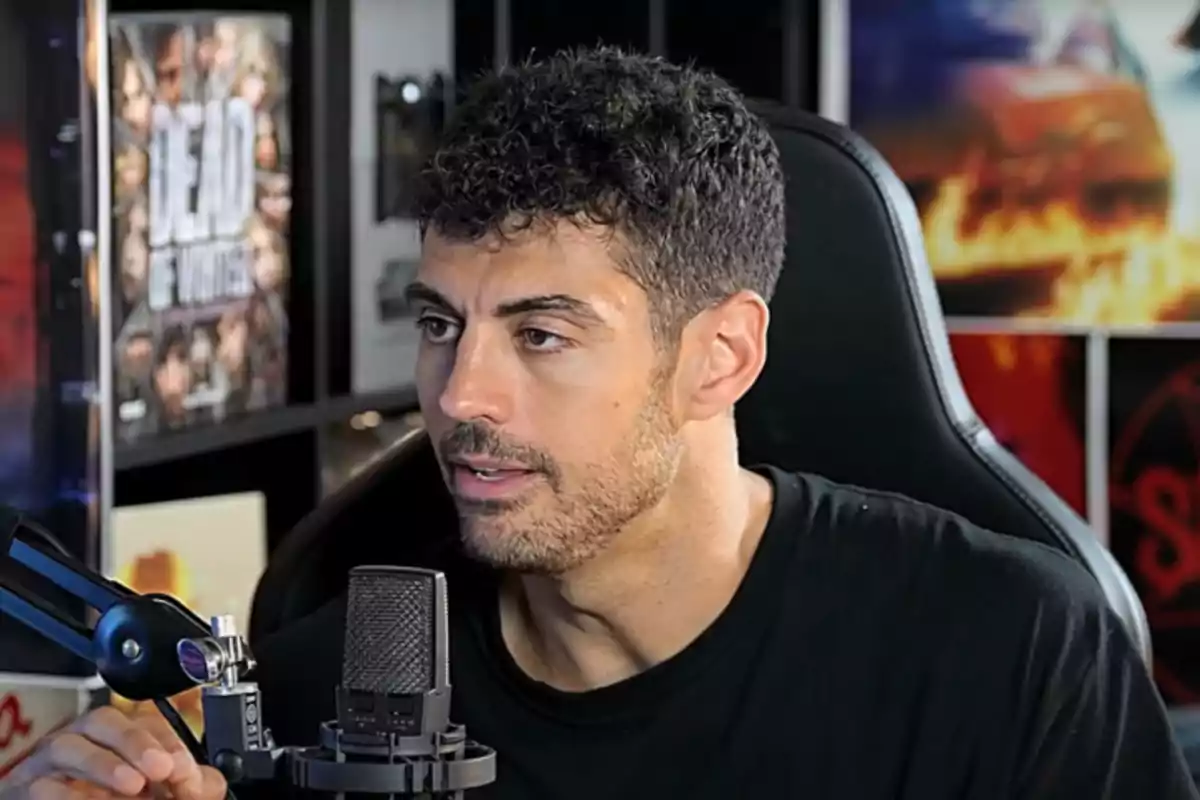 Home de cabell curt i barba retallada parlant davant d’un micròfon en un estudi amb pòsters a la paret.