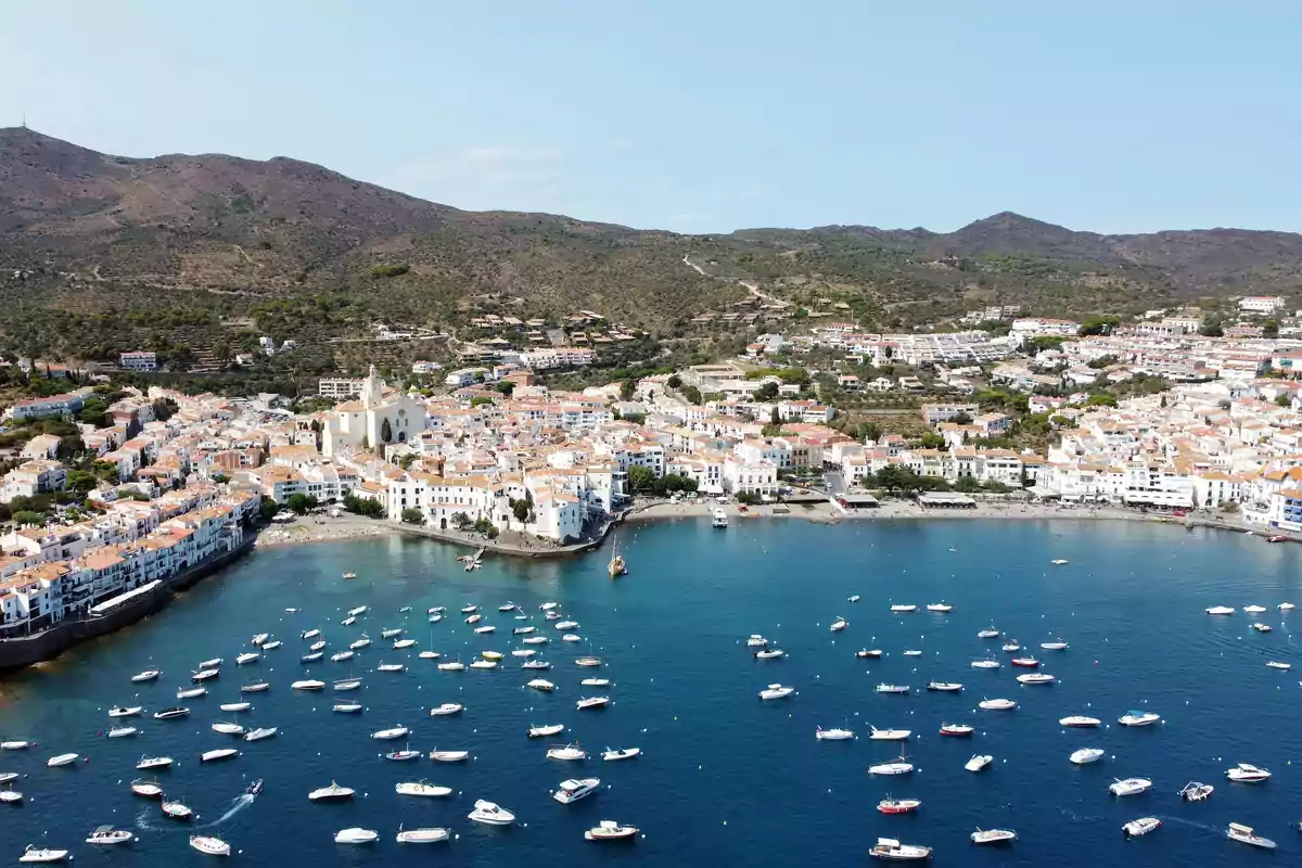 Vista aérea de un pueblo costero con casas blancas y tejados naranjas junto a una bahía llena de pequeños barcos y colinas al fondo
