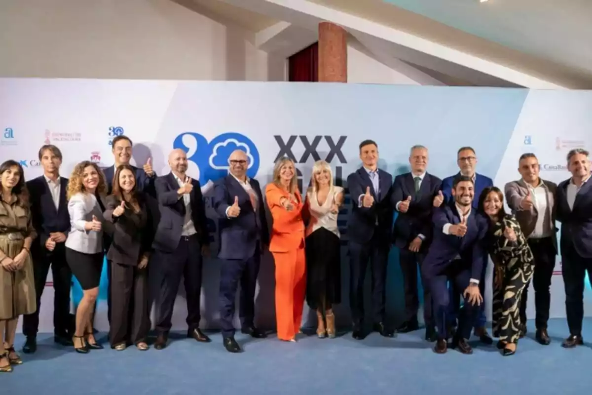 Grupo de personas vestidas de manera formal posando juntas y sonriendo mientras levantan el pulgar frente a un fondo con logotipos y el texto XXX Gala Grupo de personas vestidas de manera formal posando juntas y sonriendo mientras levantan el pulgar frente a un fondo con logotipos y el texto XXX Gala