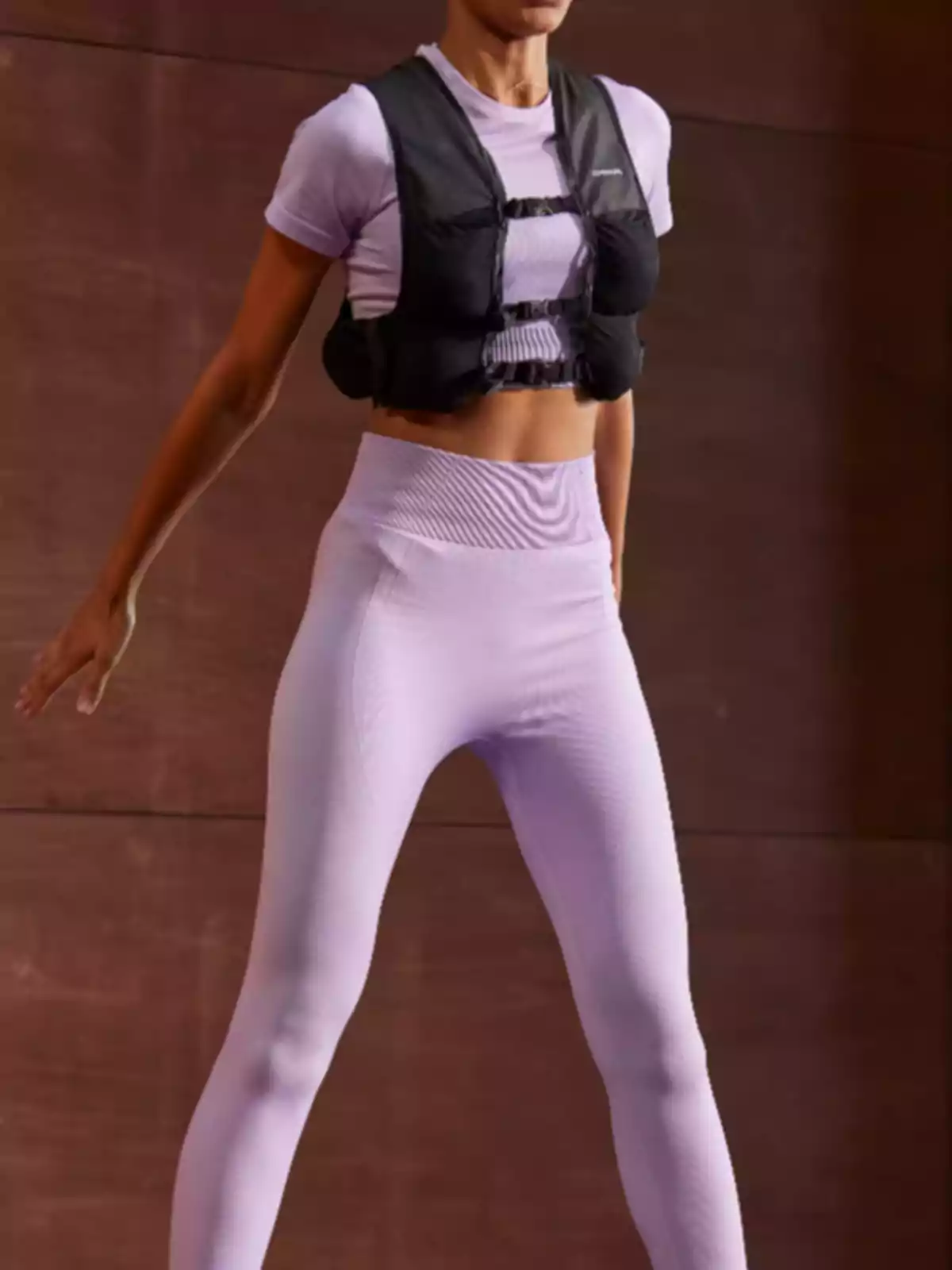 Persona amb roba esportiva lila i un xalec de pes negre mentre fa exercici en un espai interior. Persona amb roba esportiva lila i un xalec de pes negre mentre fa exercici en un espai interior.